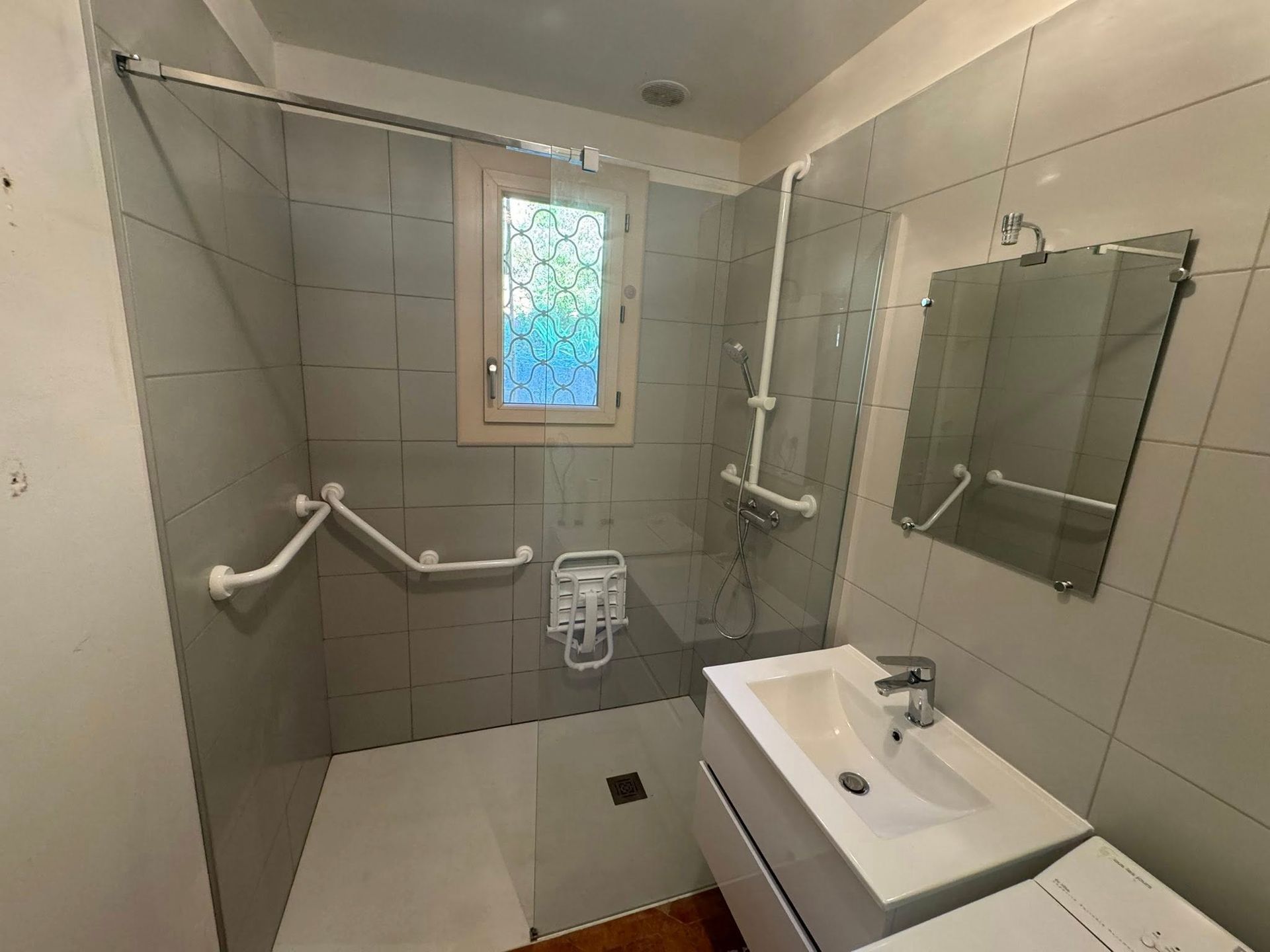Salle de bains accessible comprenant une douche à l'italienne avec des barres d'appui, un siège de douche rabattable et un lavabo mural avec miroir
