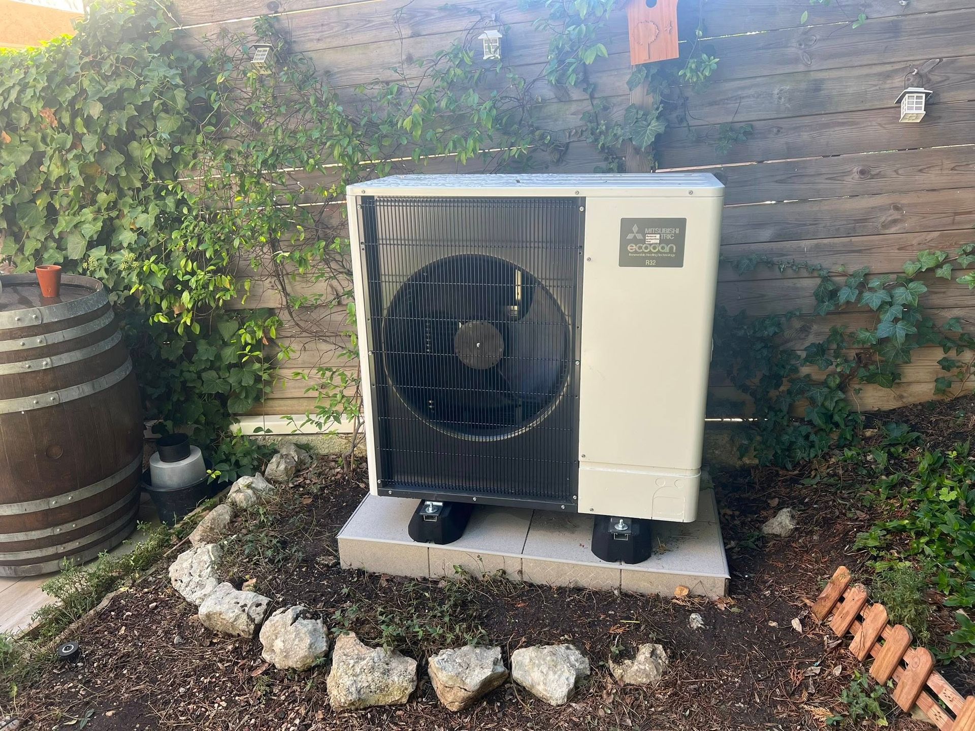Une pompe à chaleur Mitsubishi Electric est installée sur une dalle de béton dans un parterre de jardin, avec un tonneau en bois et une clôture en arrière-plan