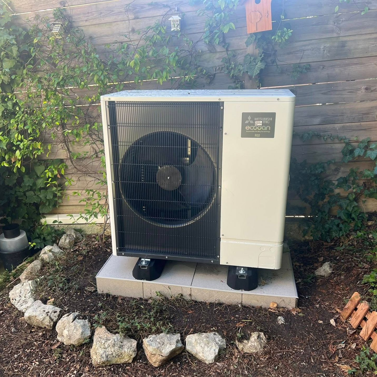 Une unité de pompe à chaleur extérieure est installée sur une dalle de béton dans un jardin, entourée de paillis, de pierres et d'une clôture en bois
