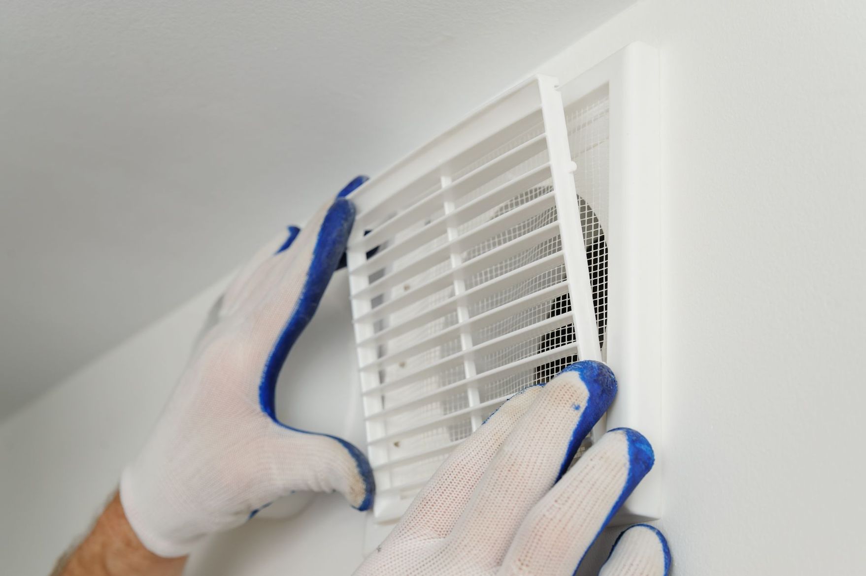 Des mains gantées de blanc avec des bordures bleues installent ou retirent une grille de ventilation rectangulaire blanche sur un mur.
