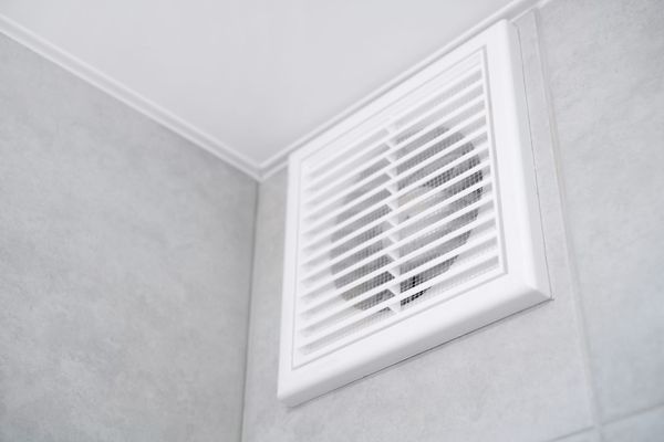Un ventilateur d'extraction de salle de bains blanc avec une grille à lamelles, fixé sur un mur carrelé gris clair dans un coin de la pièce
