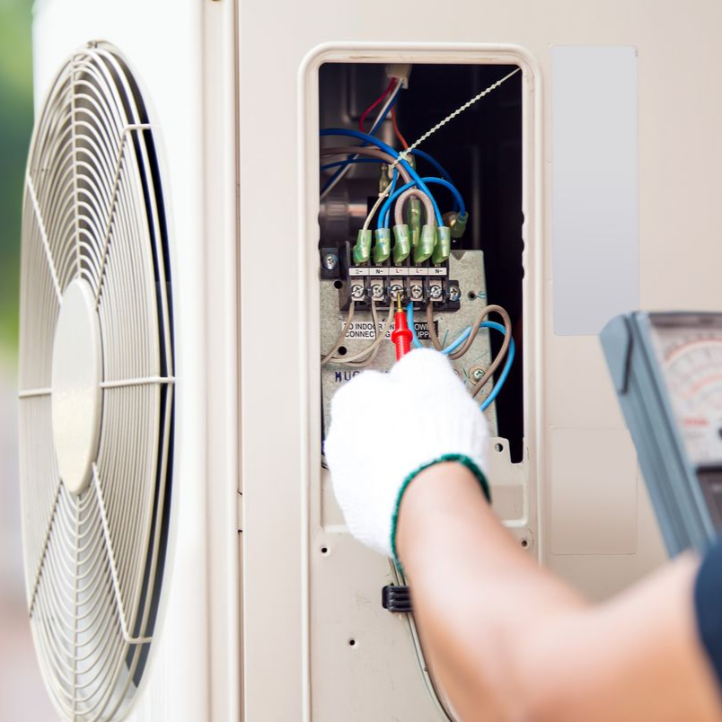 Un technicien portant des gants utilise un multimètre pour tester le câblage électrique à l'intérieur d'une unité de climatisation extérieure