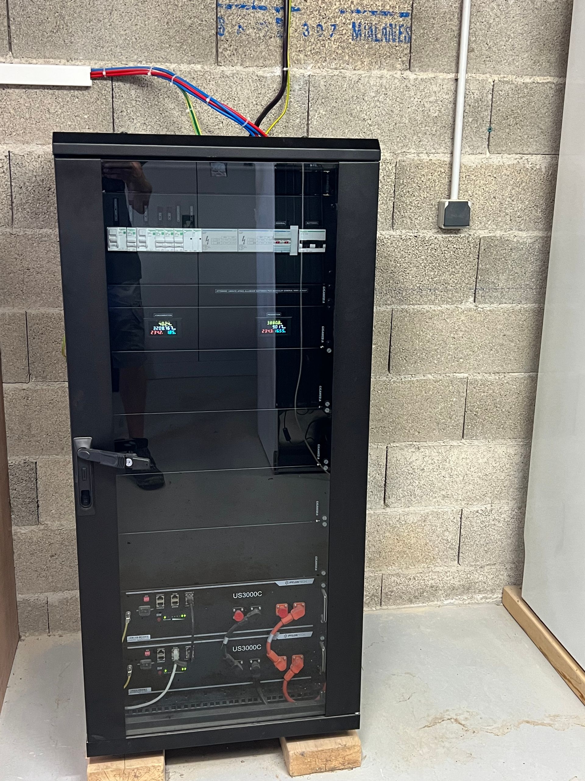 Une armoire serveur verticale noire repose sur deux petits blocs contre un mur en parpaings gris, laissant apparaître les câbles électriques au-dessus