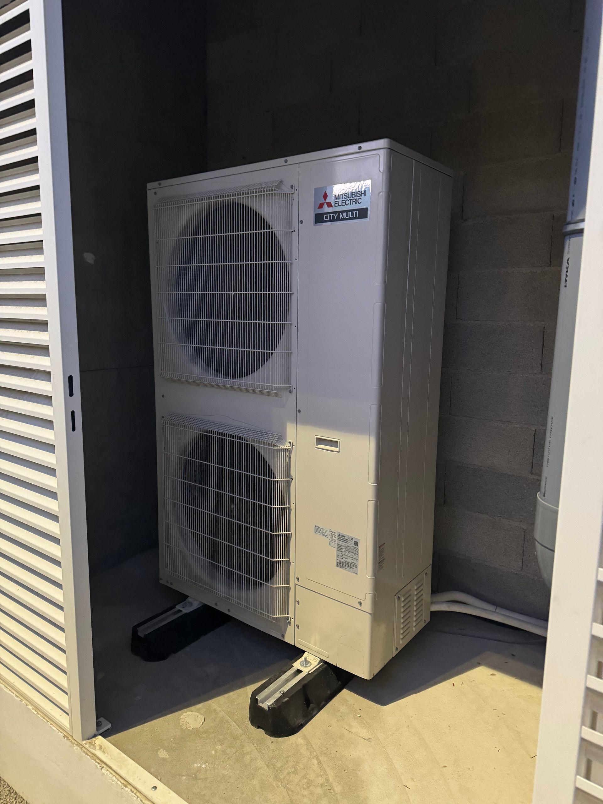 Une unité de climatisation extérieure grise repose sur des supports noirs à l'intérieur d'une alcôve en béton