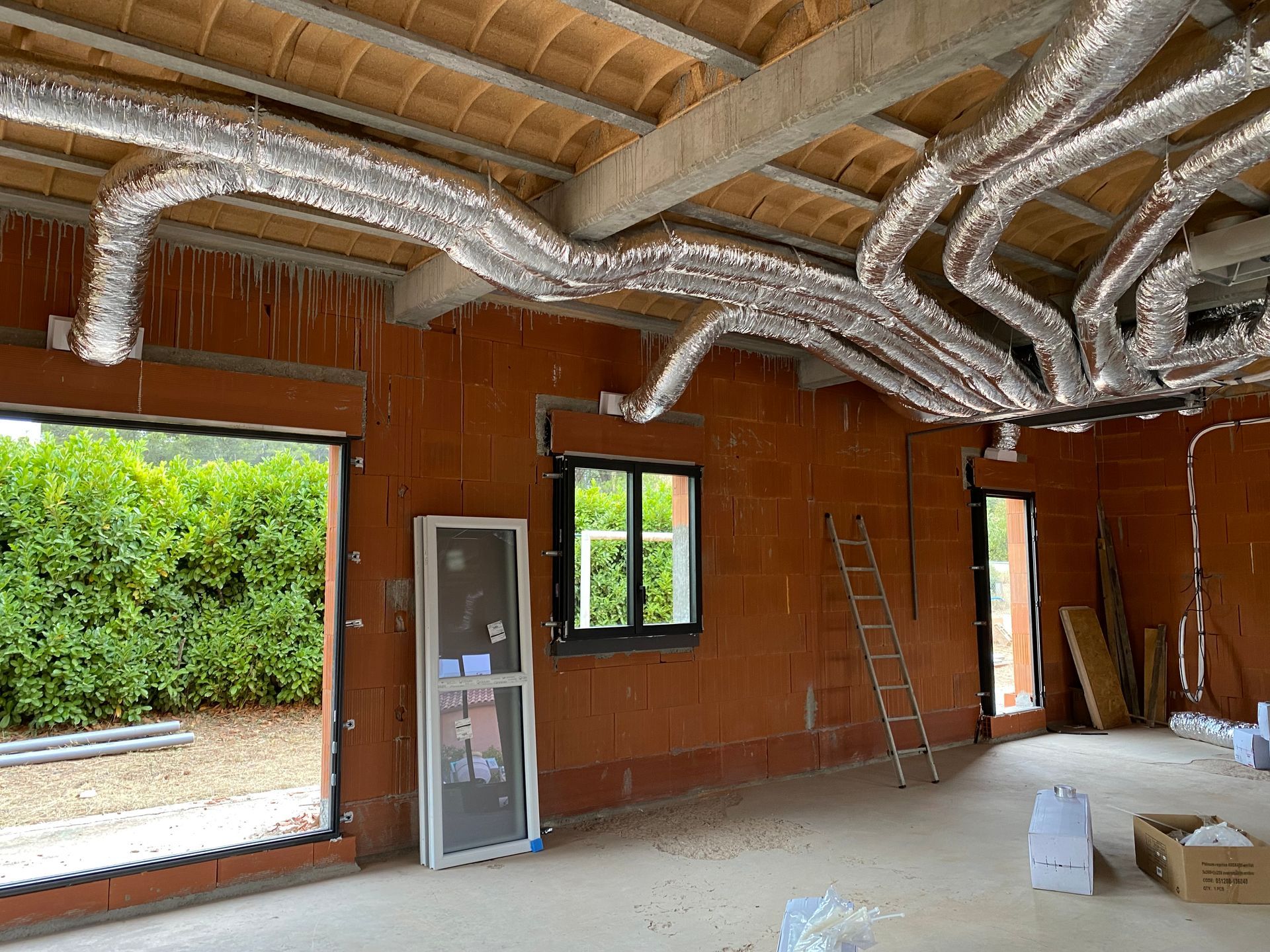 Une pièce inachevée avec des murs en briques orange, une grande fenêtre et des conduits de climatisation flexibles fixés au plafond
