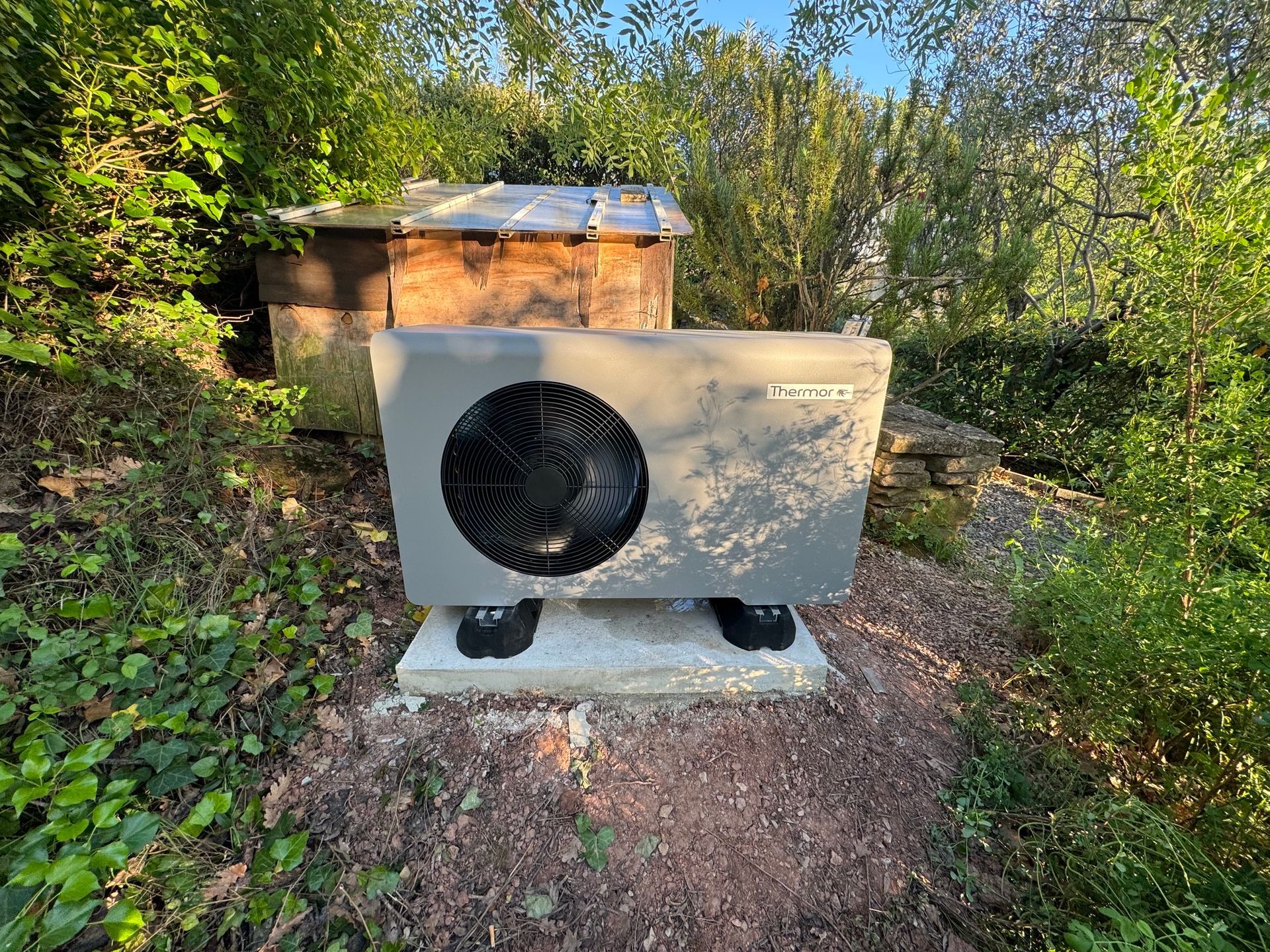 Une pompe à chaleur Thermor est installée sur une dalle de béton dans un jardin, avec un cabanon en bois en arrière-plan