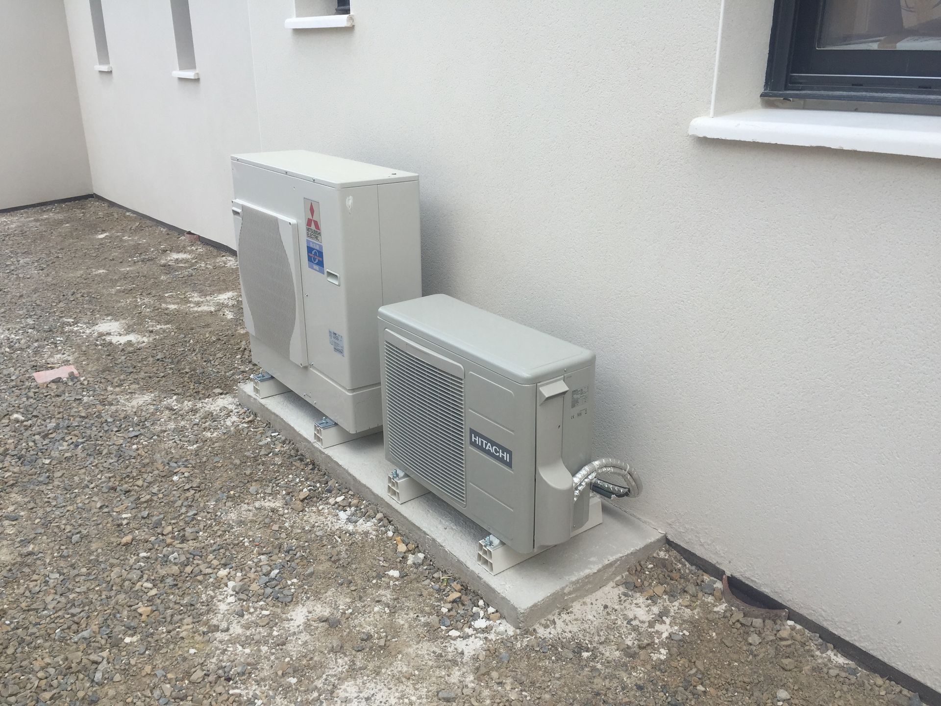 Deux unités de pompe à chaleur CVC gris clair sont installées sur une dalle de béton contre le mur extérieur d'un bâtiment