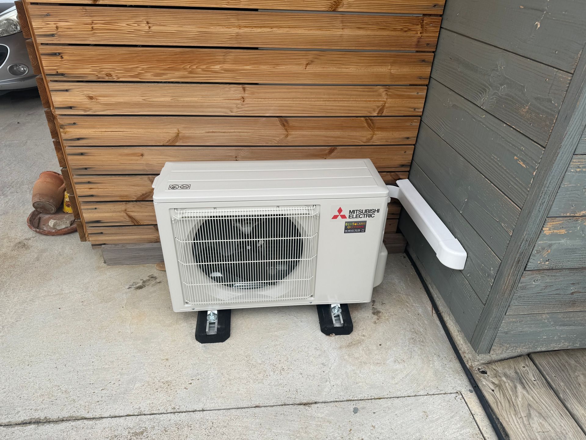 Un climatiseur extérieur Mitsubishi beige repose sur des supports en caoutchouc noir, sur une terrasse en béton, à côté de murs en bois