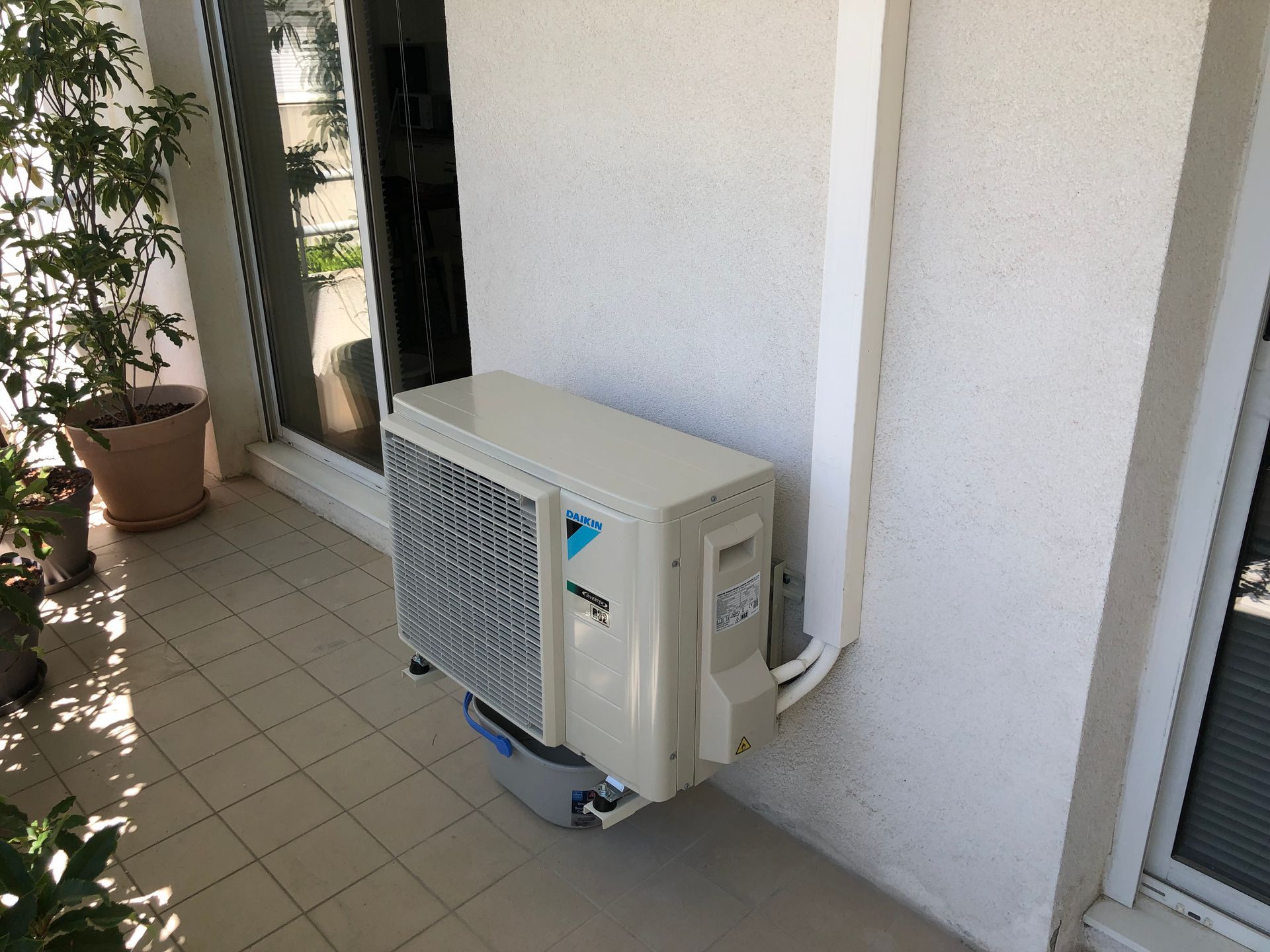 Un climatiseur extérieur Daikin beige est installé sur un balcon contre un mur blanc, à côté d'une porte vitrée