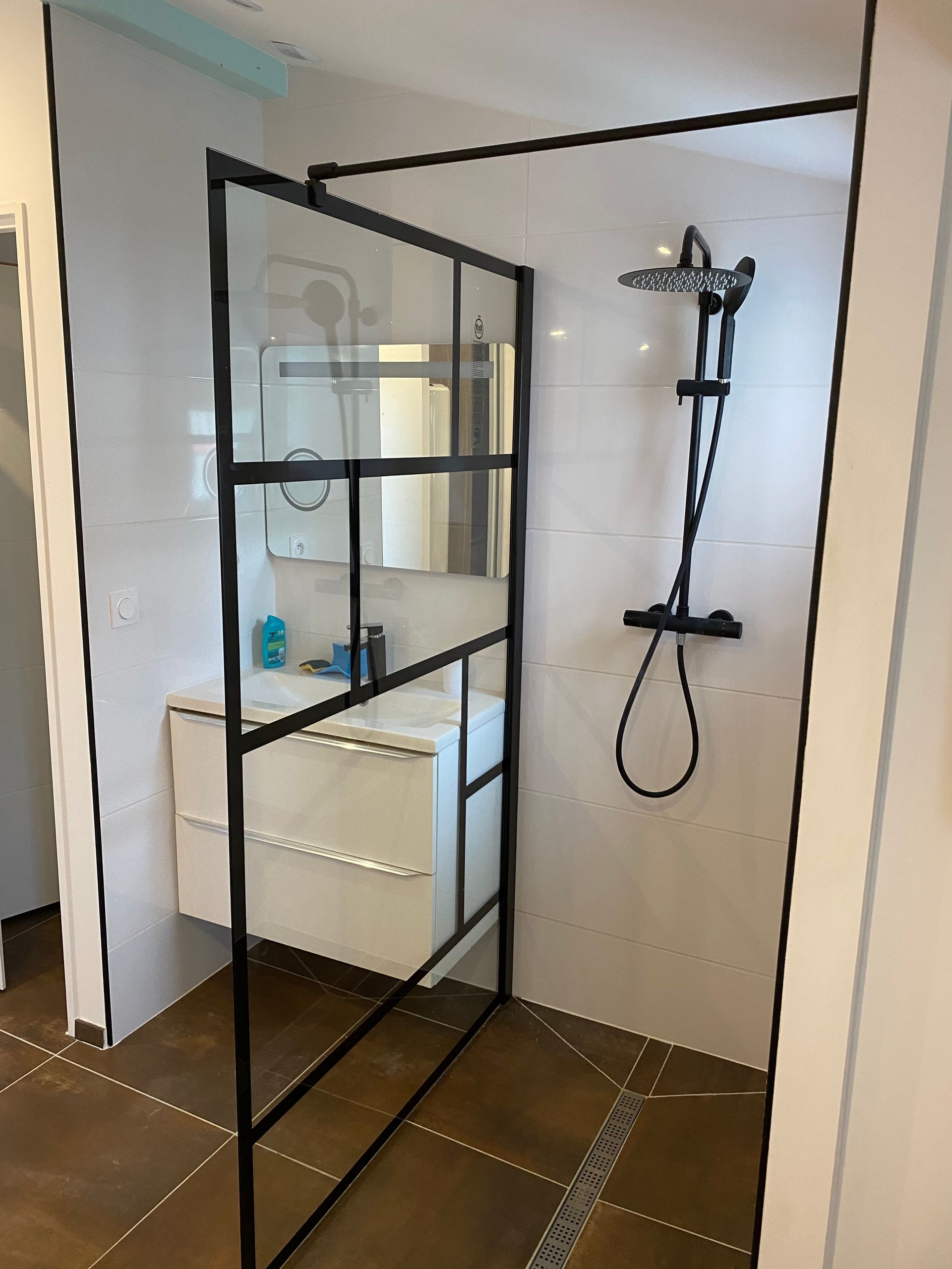 Un espace douche moderne avec une paroi de douche en verre à cadre noir, des accessoires de douche noir mat et un sol carrelé marron
