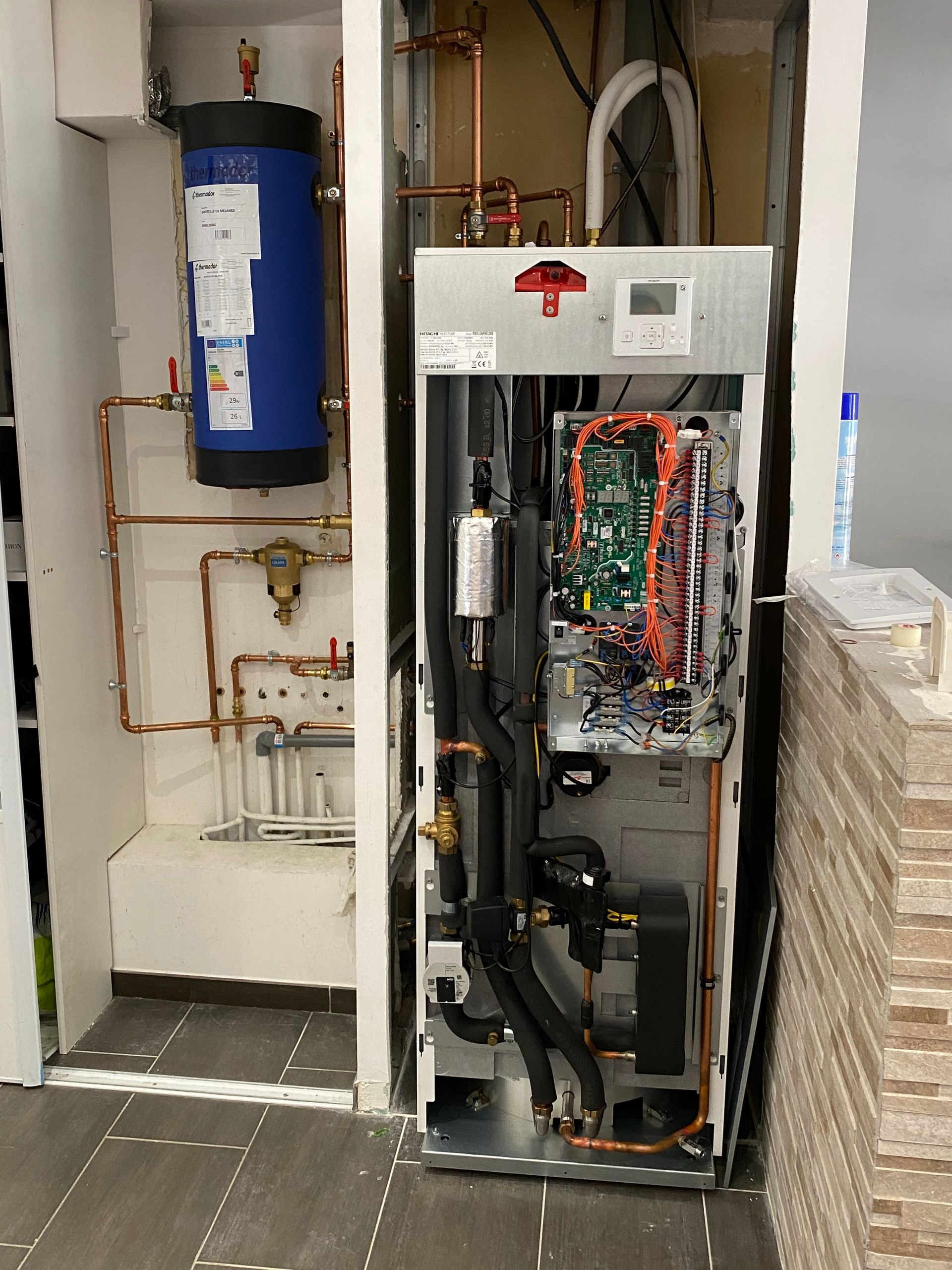 Un ballon d'eau chaude sanitaire bleu à côté d'une unité de pompe à chaleur ouverte de style industriel avec des composants internes apparents