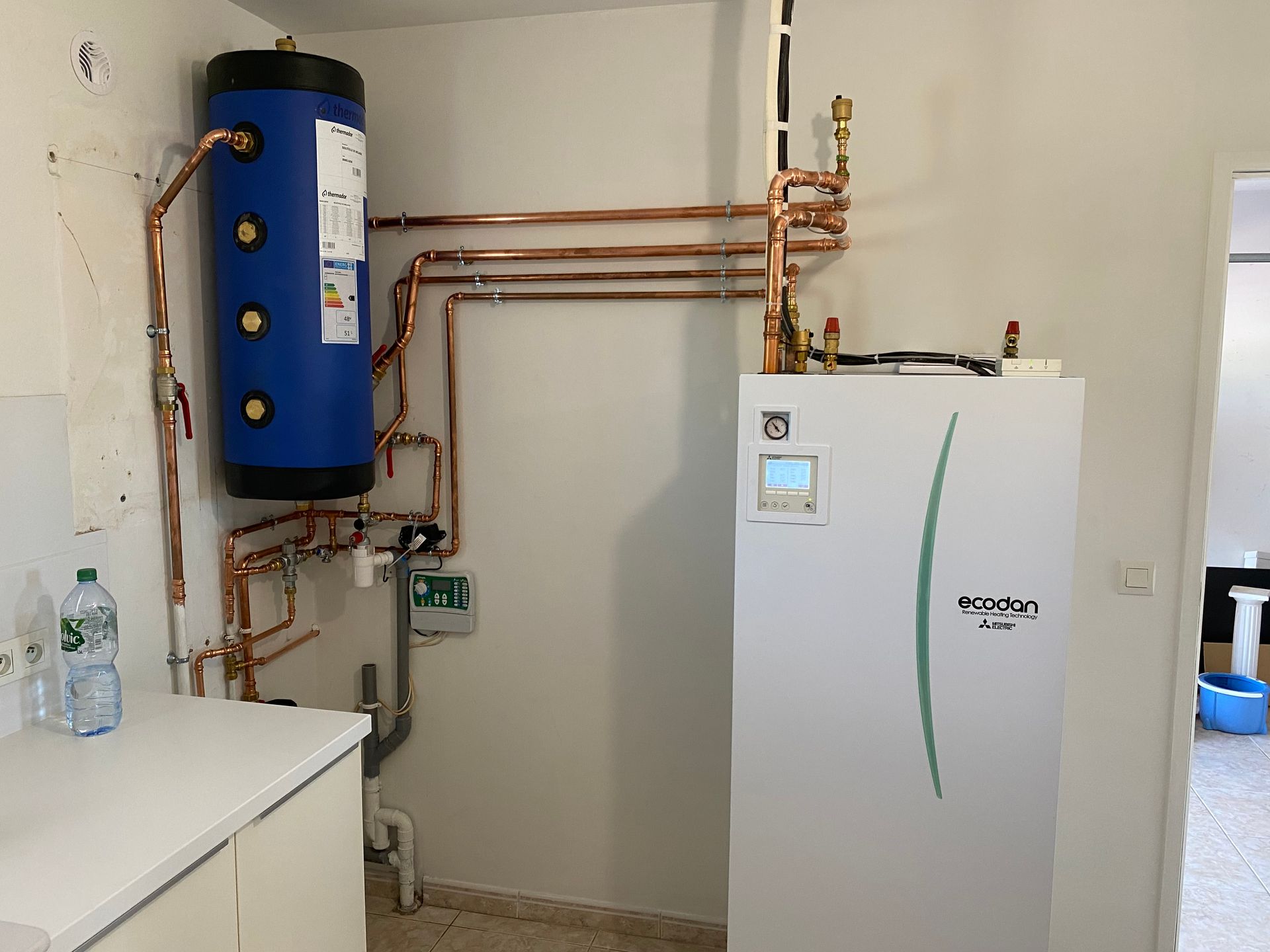 Un chauffe-eau bleu et une grande chaudière blanche reliés par des tuyaux en cuivre dans une buanderie