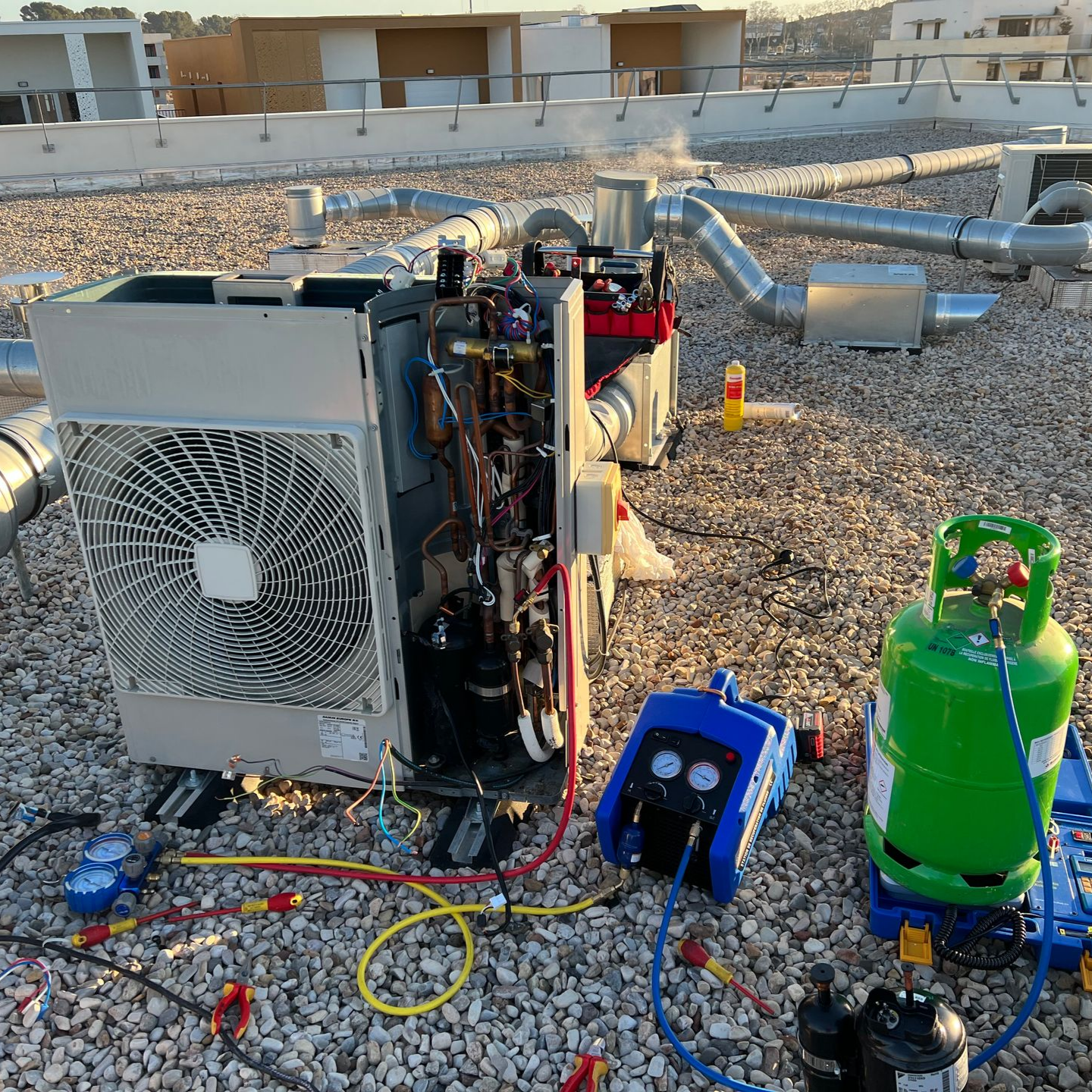 Outils de technicien reliés à une unité de climatisation sur le toit, avec un réservoir de réfrigérant vert et des manomètres pour la maintenance