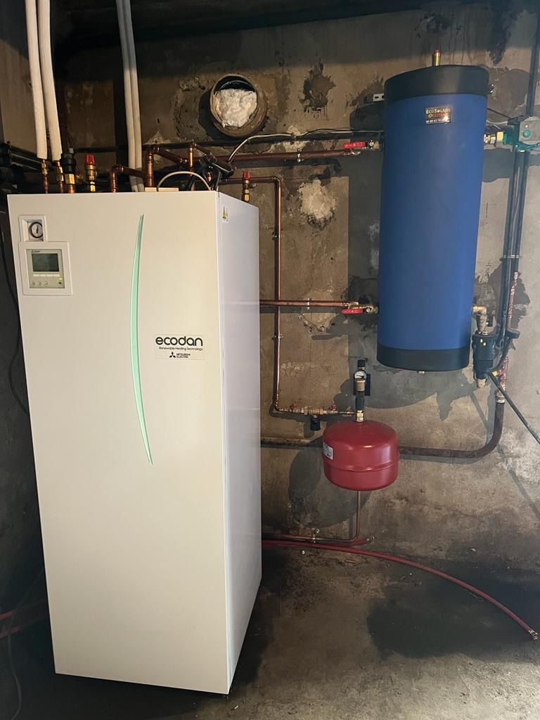 Dans une buanderie, un appareil de chauffage Viesmann blanc et un réservoir d'eau cylindrique vertical bleu, reliés par des tuyaux en cuivre
