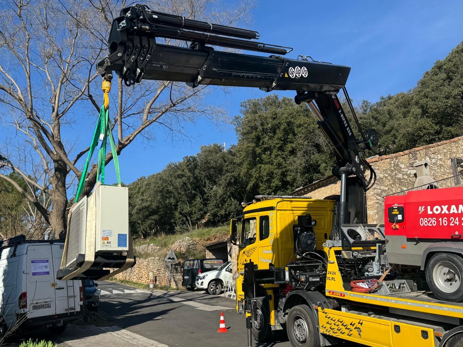 Un camion-grue jaune soulève une grande unité industrielle blanche à l'aide de sangles vertes