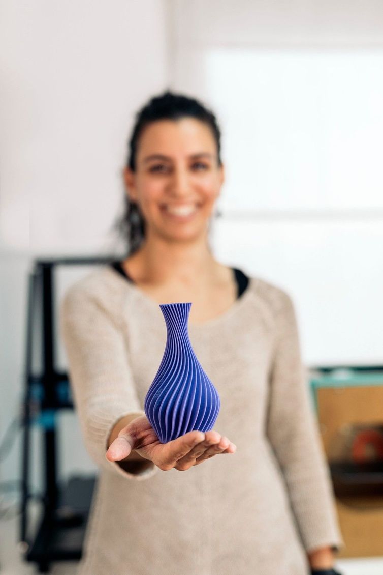 Mujer sosteniendo un jarrón impreso en 3D azul y blanco, sonriendo, con una impresora 3D en el fondo.