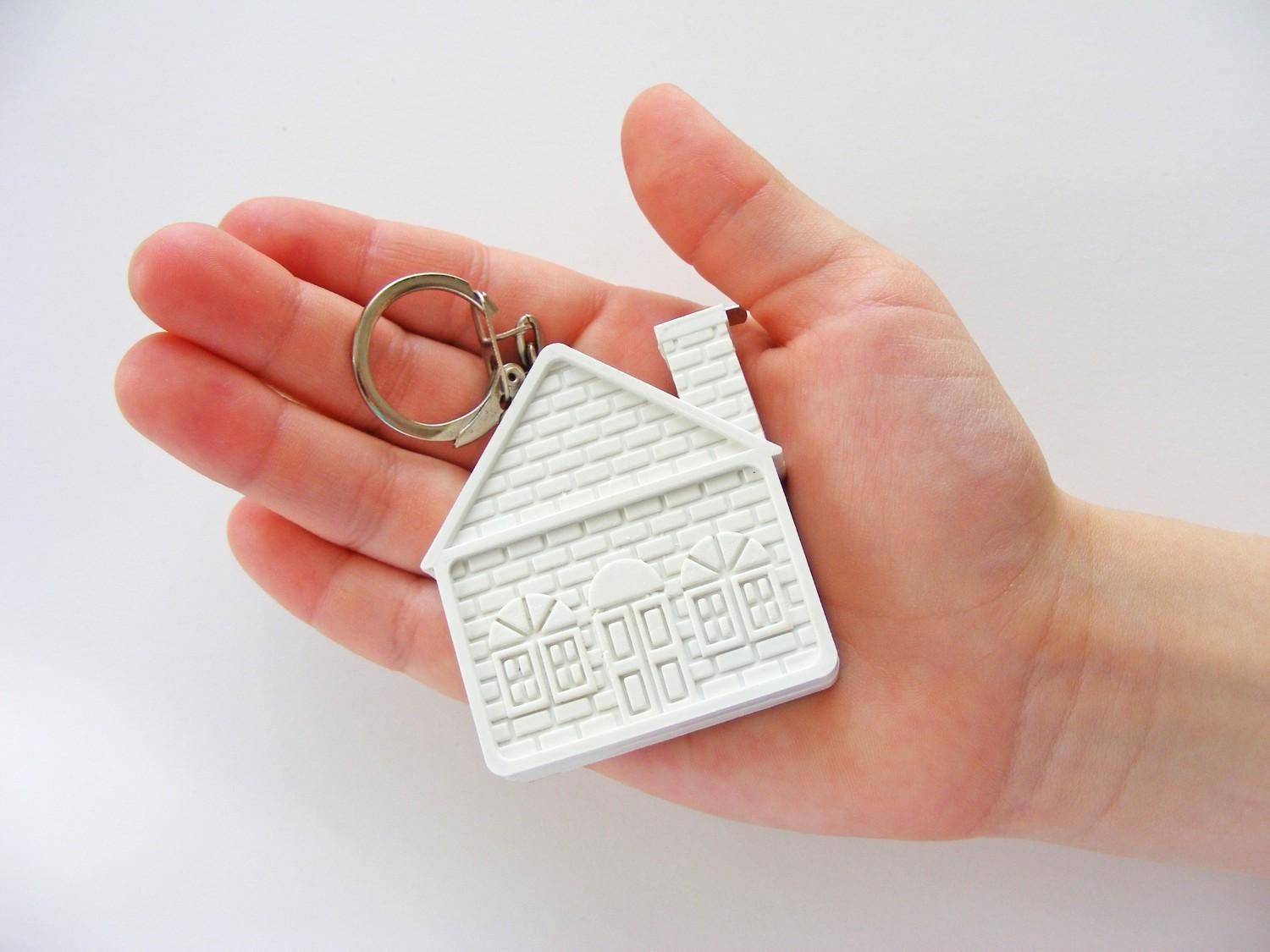 Mano sosteniendo un llavero con forma de casa blanca sobre un fondo blanco.