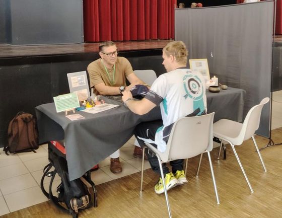 Deux personnes discutent autour d'une table sur un stand de foire, avec des panneaux et une nappe grise.
