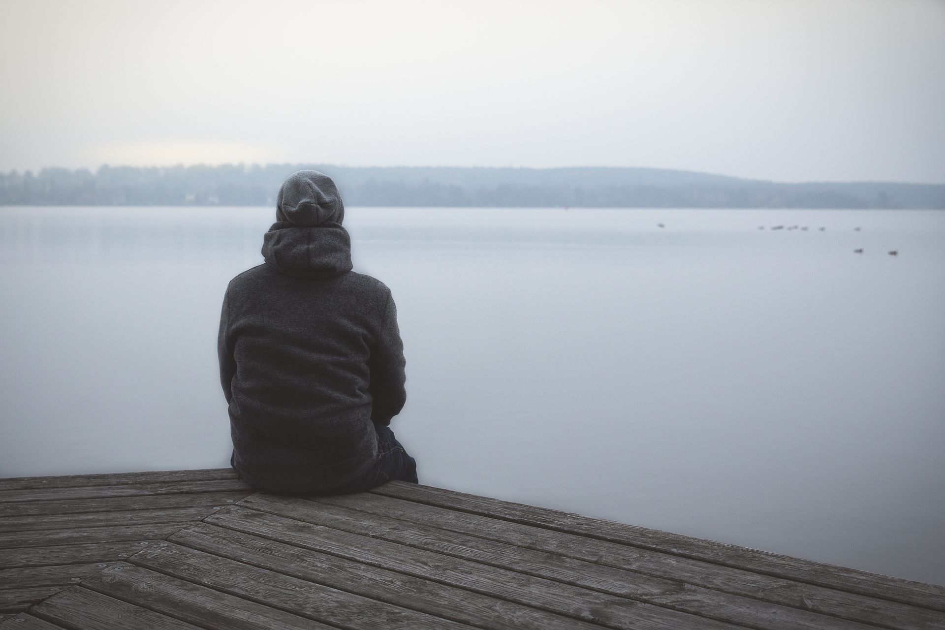 Une personne vêtue d'une veste sombre est assise sur un quai en bois, contemplant un lac calme et brumeux.