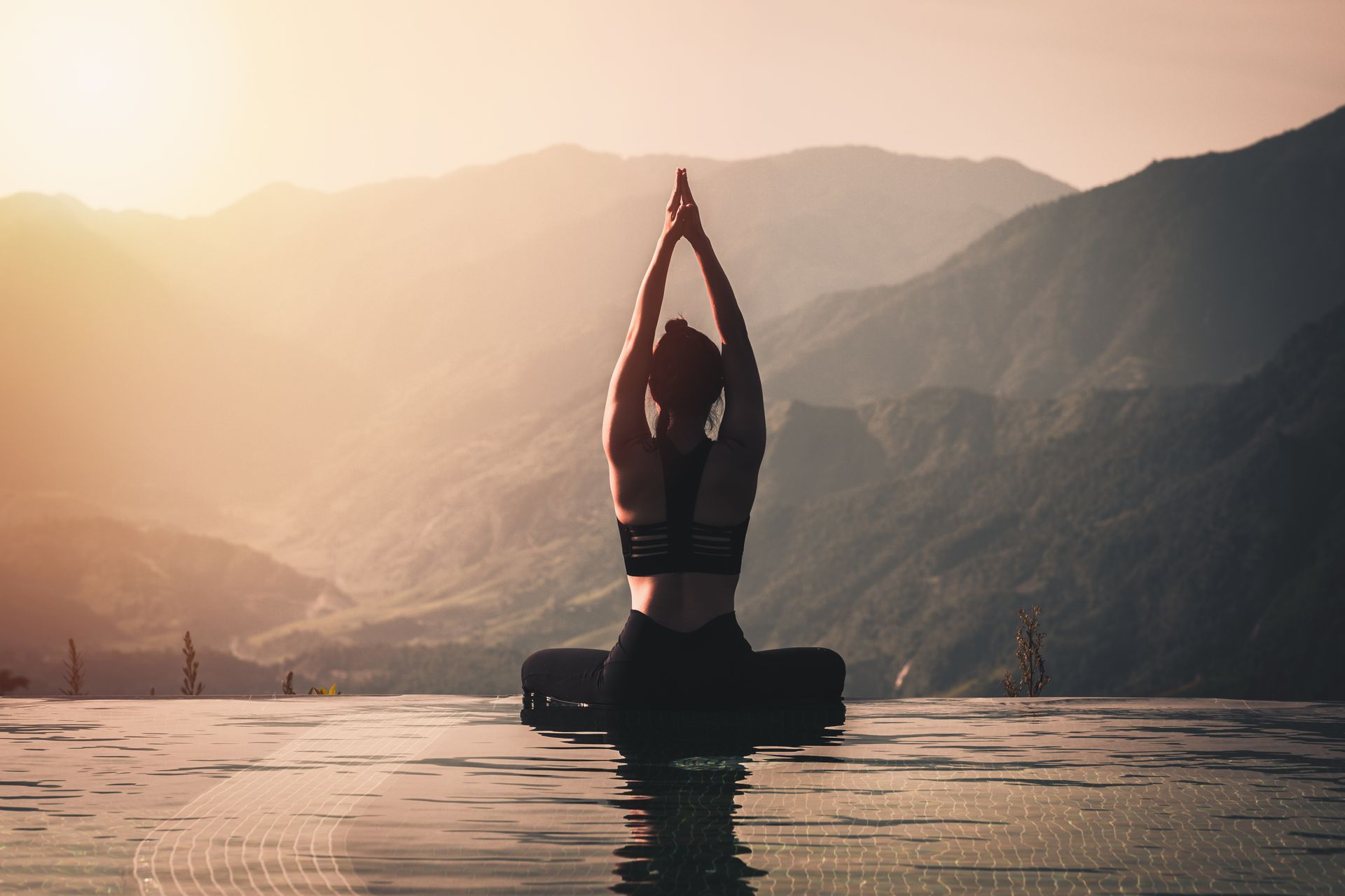 Une personne pratique le yoga dans une eau calme au lever du soleil, avec des montagnes en arrière-plan.
