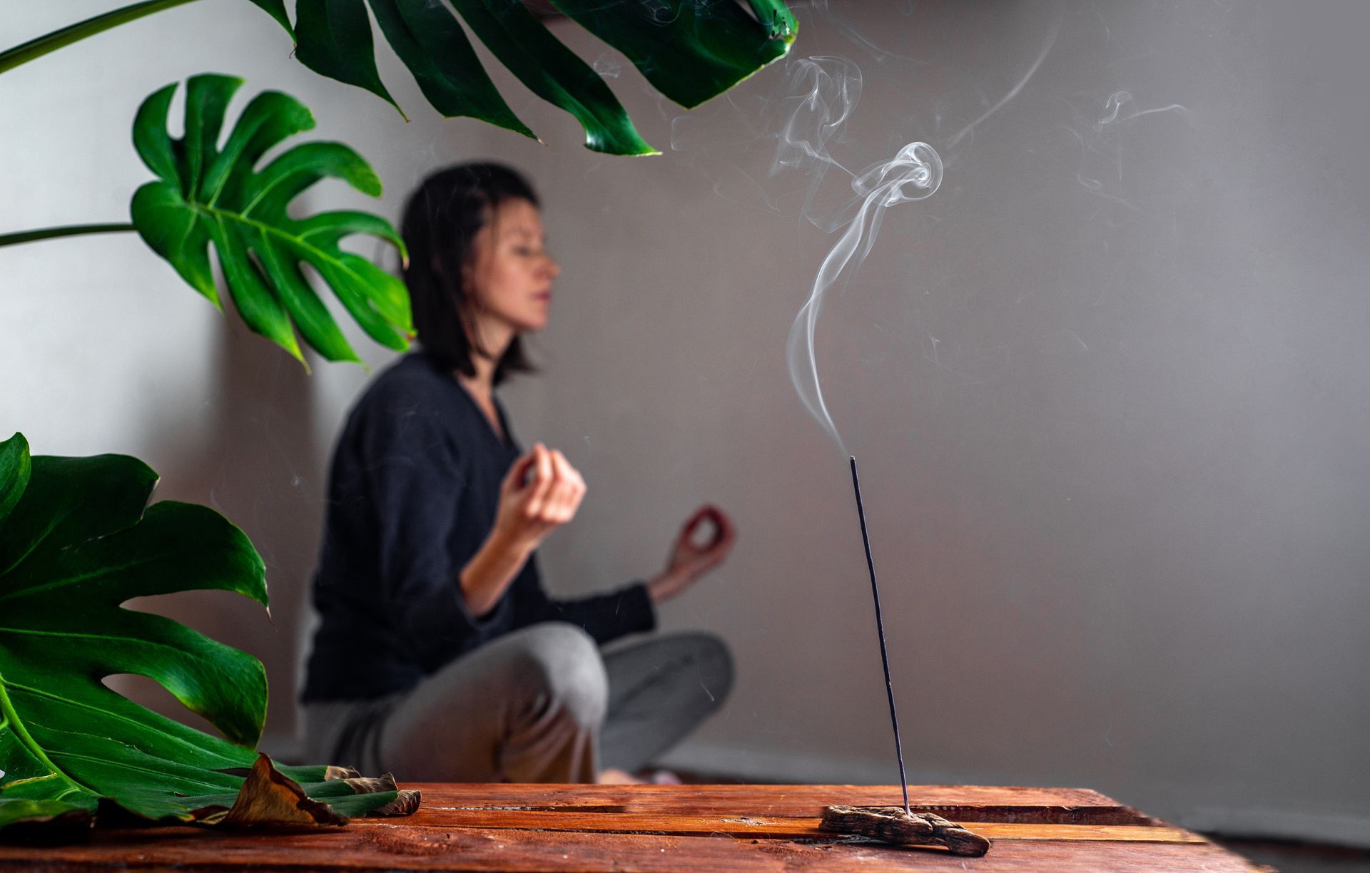 Une personne médite à l'intérieur, près d'un bâtonnet d'encens allumé, avec de grandes feuilles vertes au premier plan.