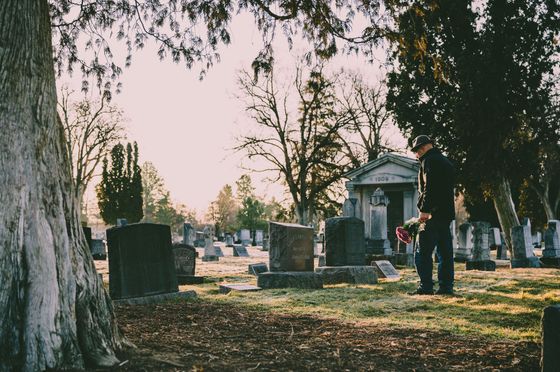 Une personne vêtue de noir se tient dans un cimetière, à côté d'un petit mausolée, au crépuscule.