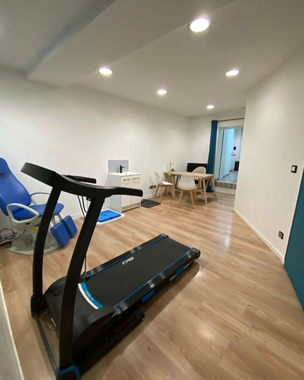 Salon lumineux avec tapis de course, fauteuil bleu, table à manger et spots encastrés au plafond