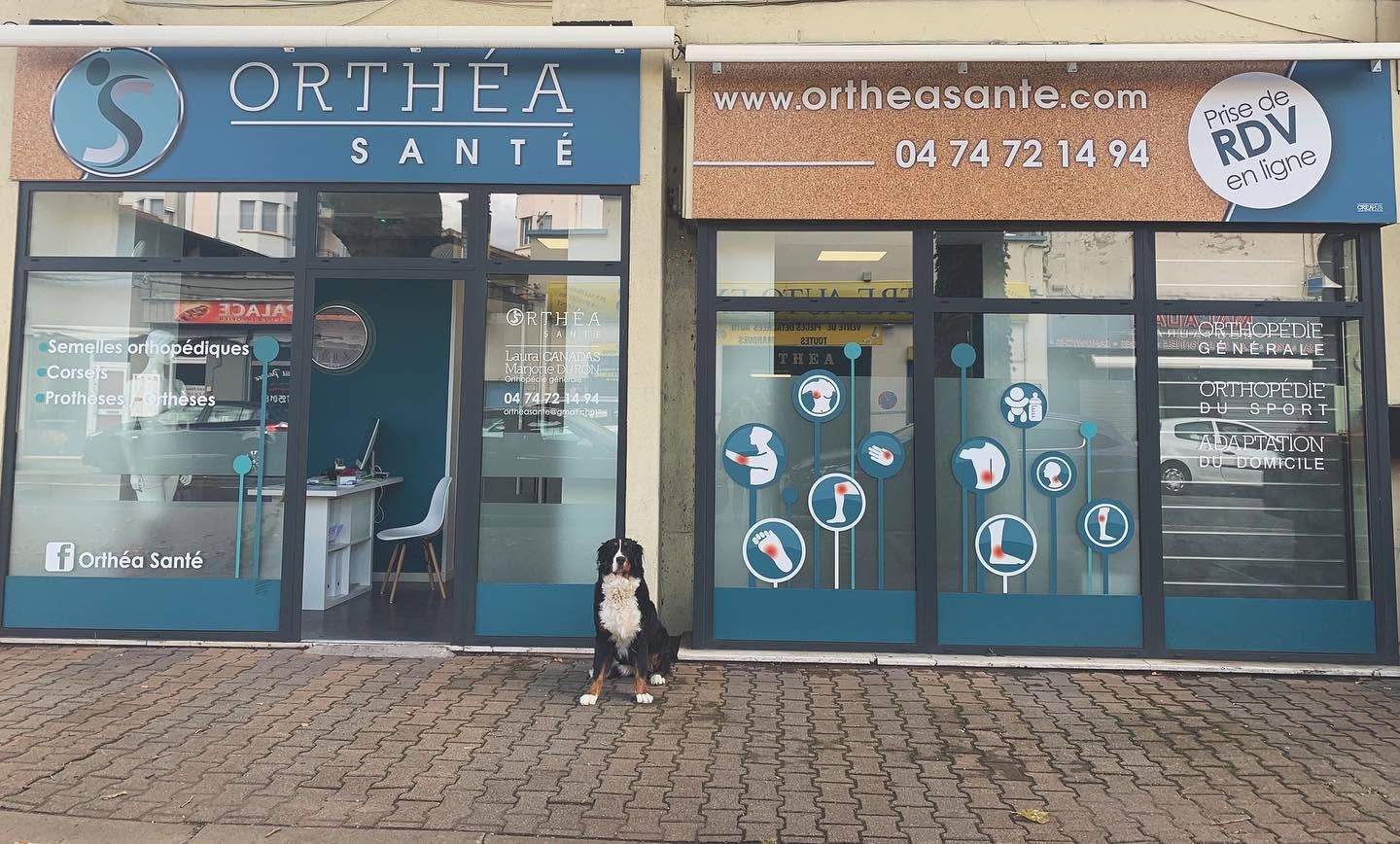 Devanture du cabinet avec enseigne bleue Orthéa Santé, un chien pose devant la vitrine