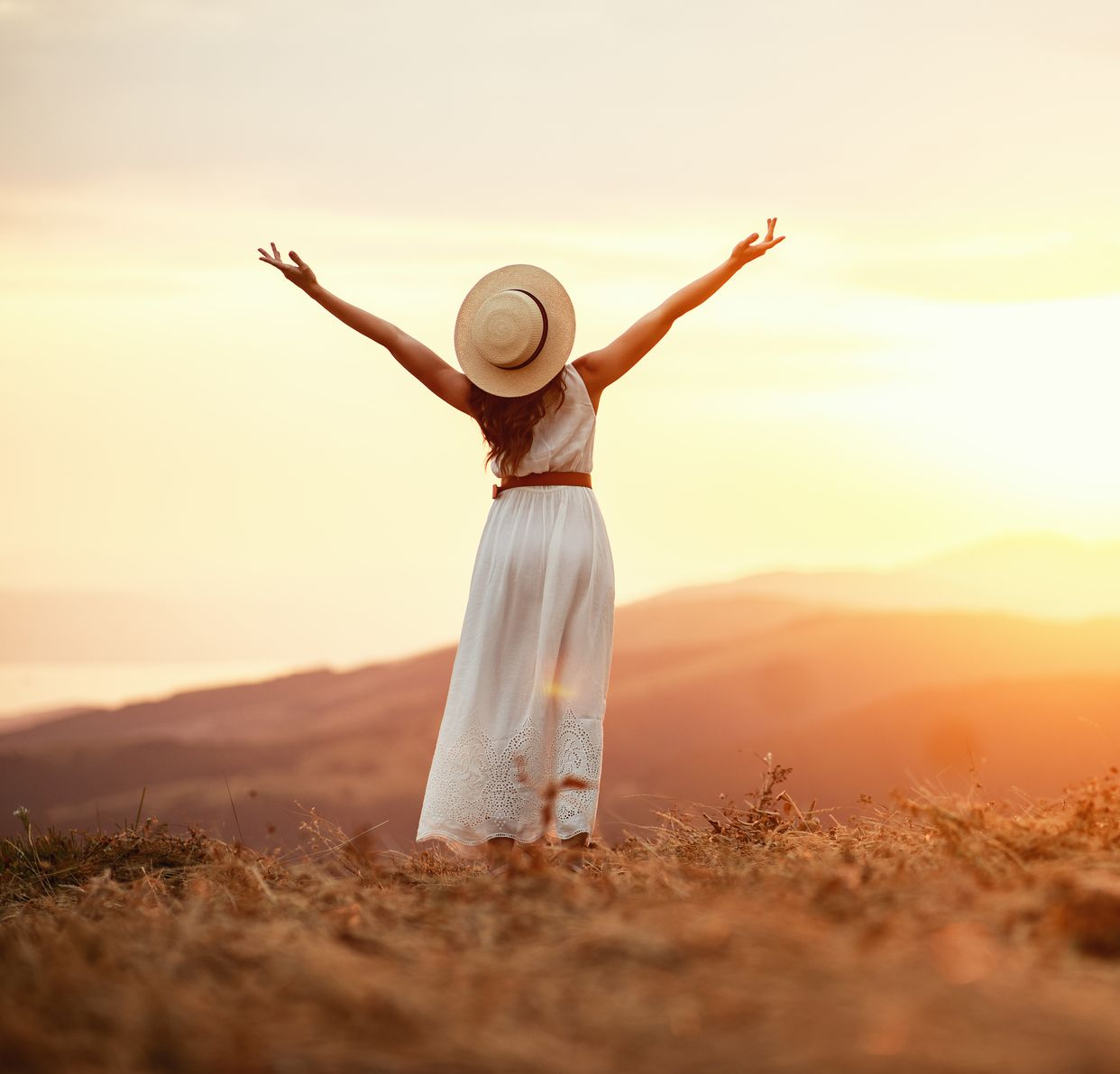 Une femme en robe et chapeau blancs se tient debout, les bras levés, dans un champ ensoleillé au coucher du soleil.