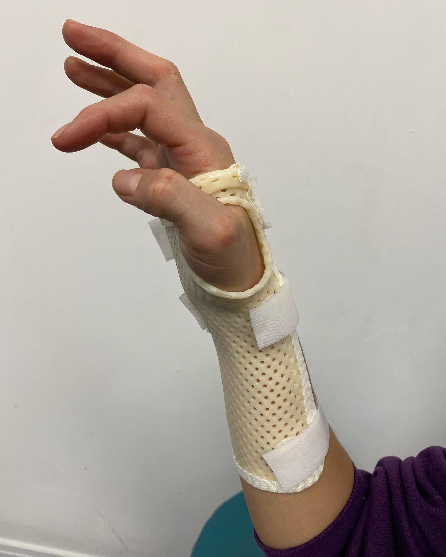 Main portant une attelle de poignet blanche micro perforée avec support pour le pouce, appuyée contre un mur nu.
