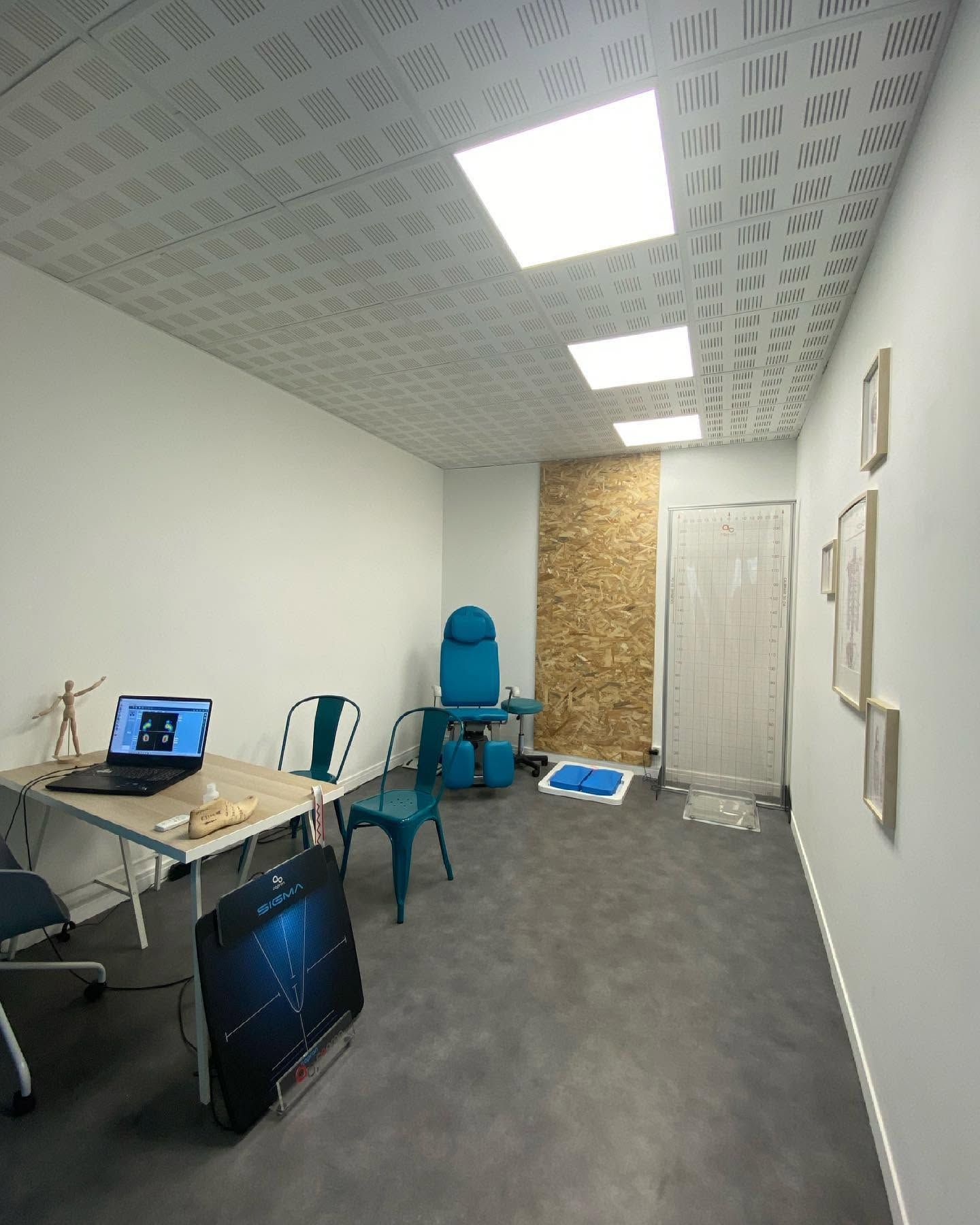 Petit bureau avec bureau, ordinateur portable, chaise bleue, moquette grise, murs blancs et un tableau en liège au fond.