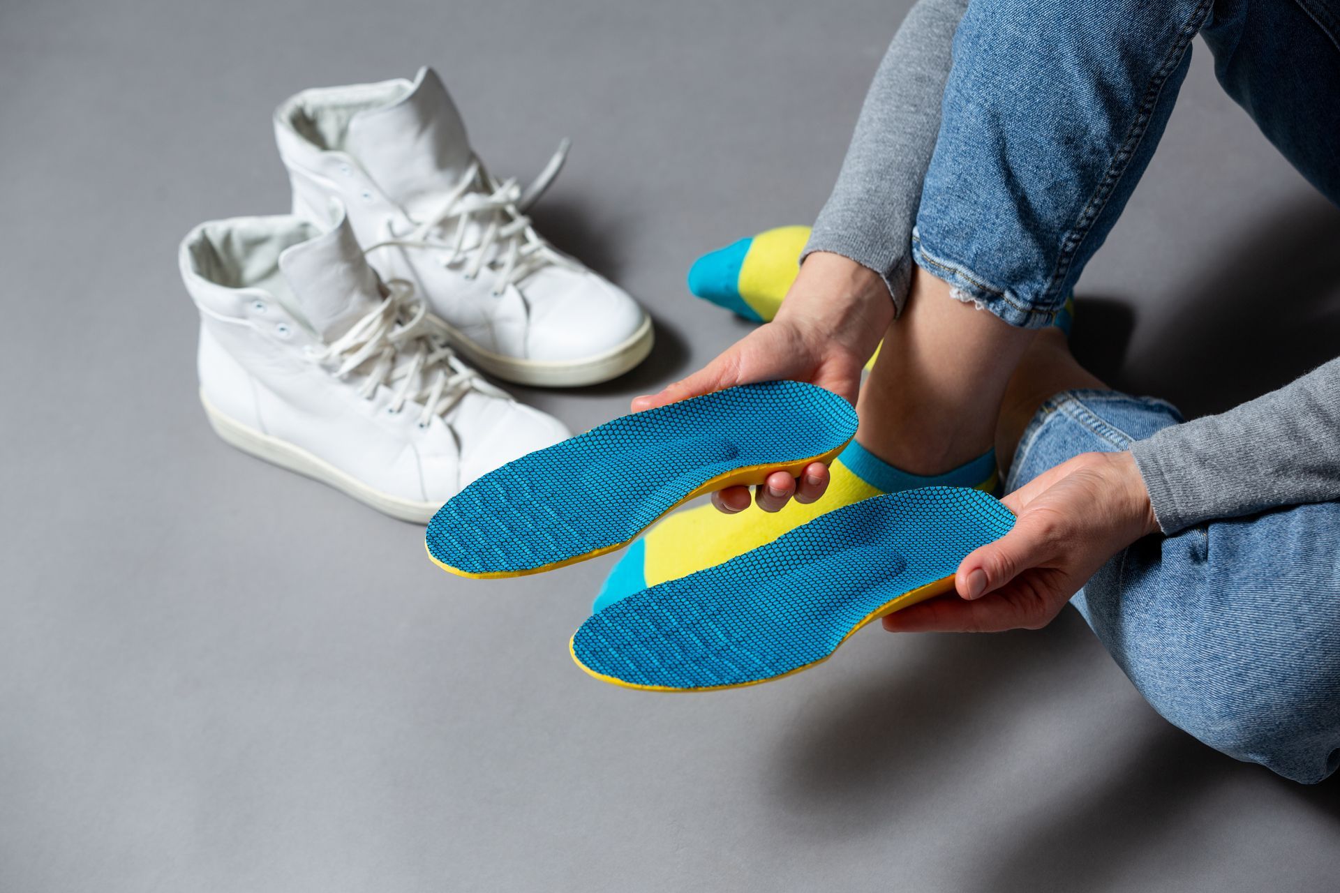 Une personne tenant des semelles bleues et jaunes à côté de baskets blanches sur un sol gris
