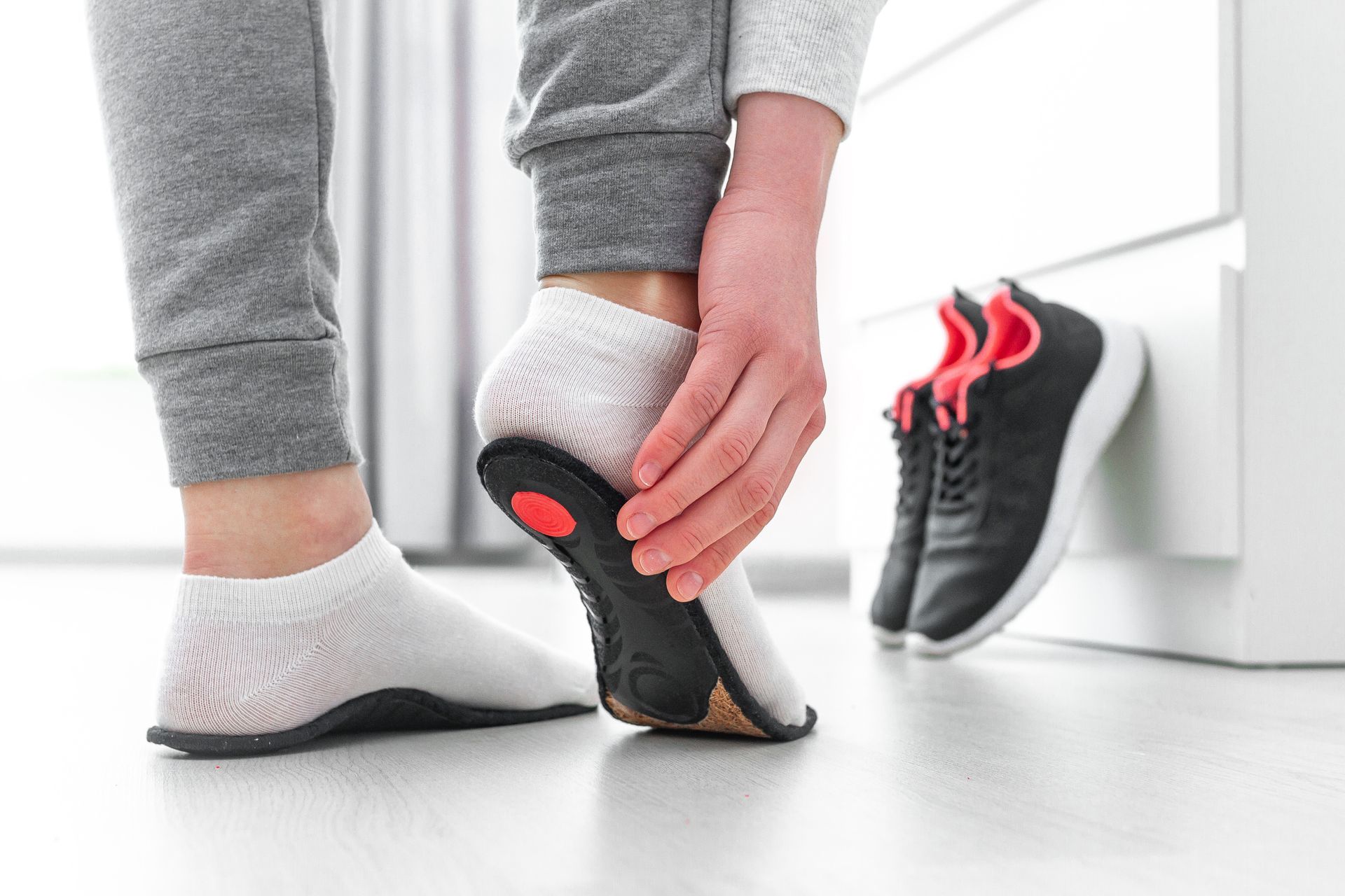 Une personne se tient le talon, son pied droit est posé sur la semelle de ses baskets. Ces dernières sont à proximité.