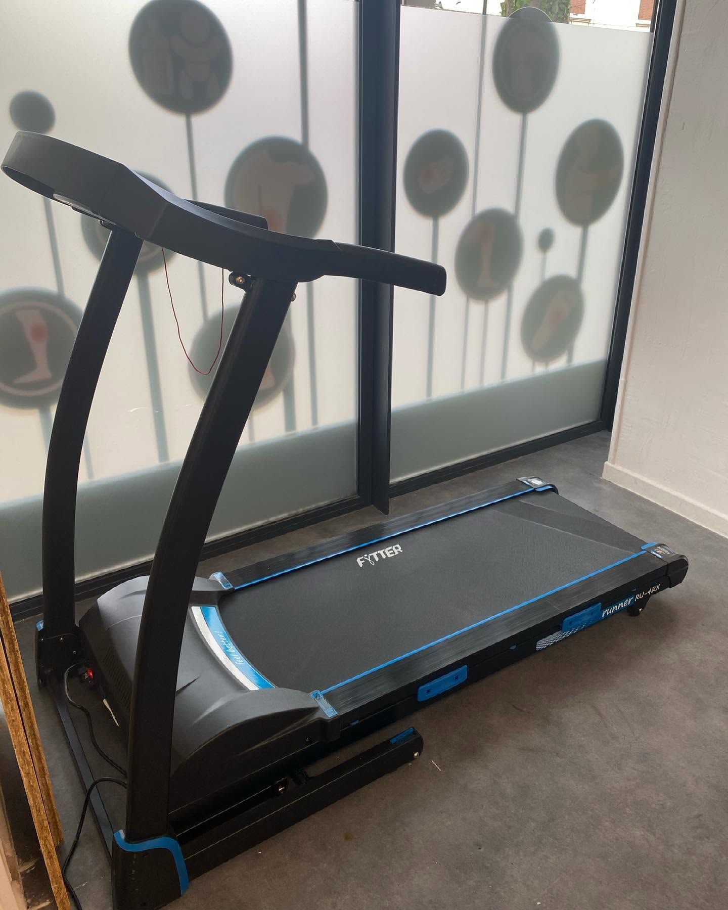 Tapis roulant noir dans une salle de sport, vu de côté près d'une paroi en verre dépoli.