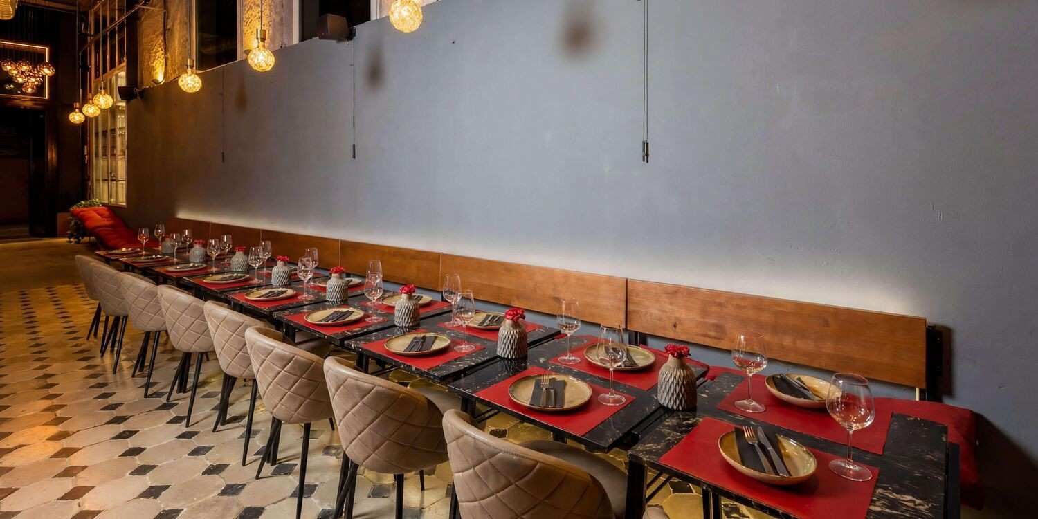 Una larga mesa de banquete con manteles individuales y platos rojos se extiende a lo largo de un comedor con paredes grises.