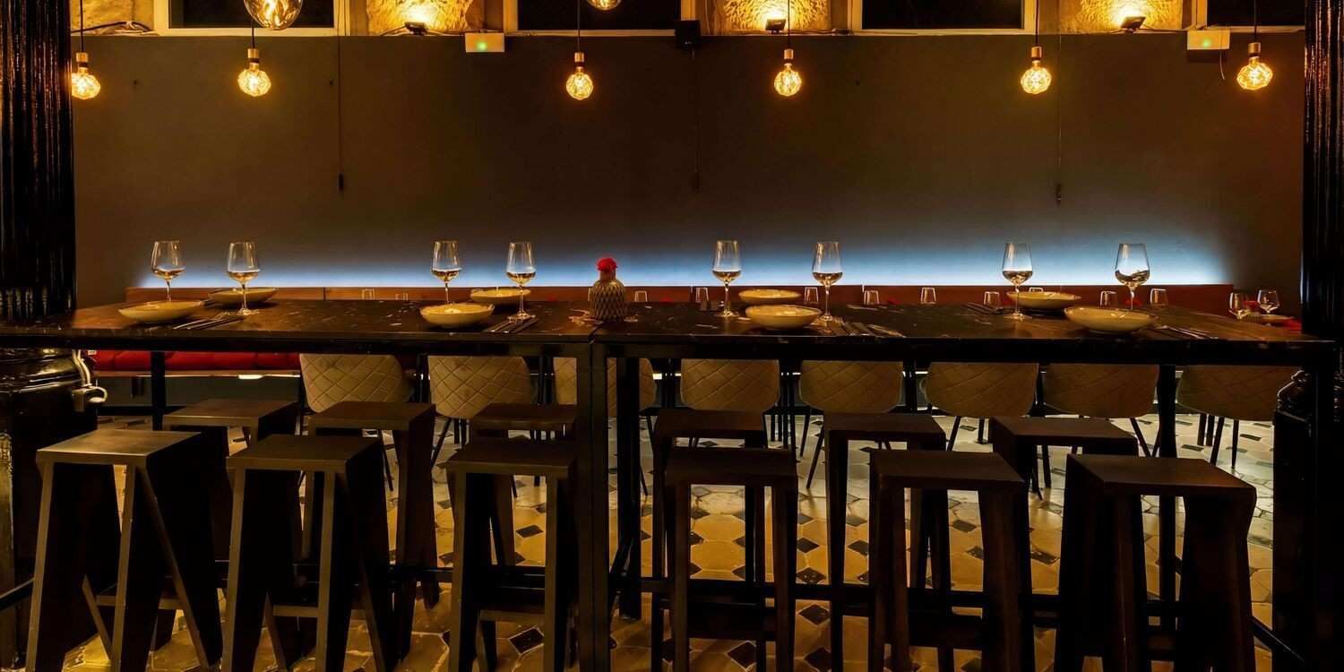 Bar con iluminación tenue, taburetes, una larga barra y luces colgantes cálidas sobre un interior de madera.