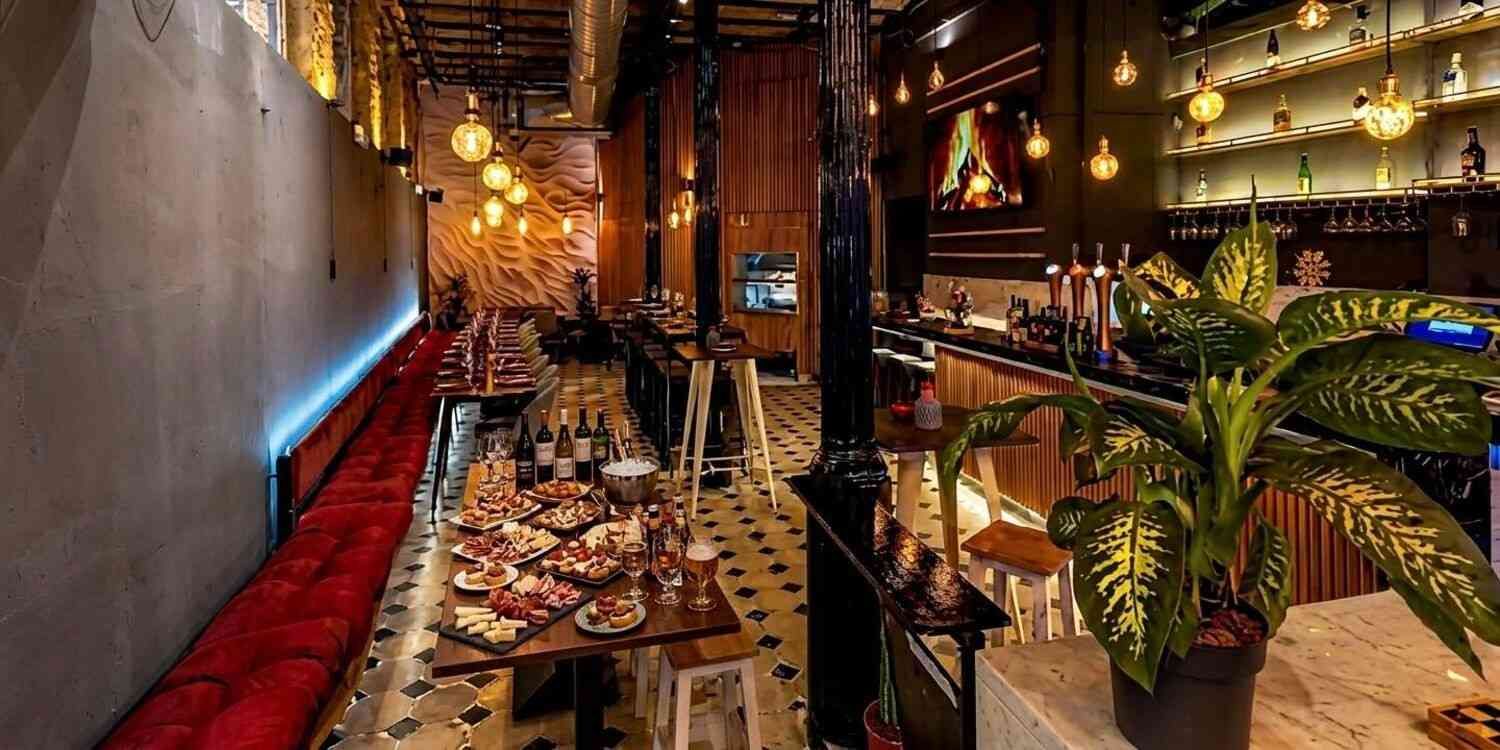 Interior del restaurante con iluminación tenue, largas mesas comunitarias, lámparas colgantes cálidas y plantas exuberantes.