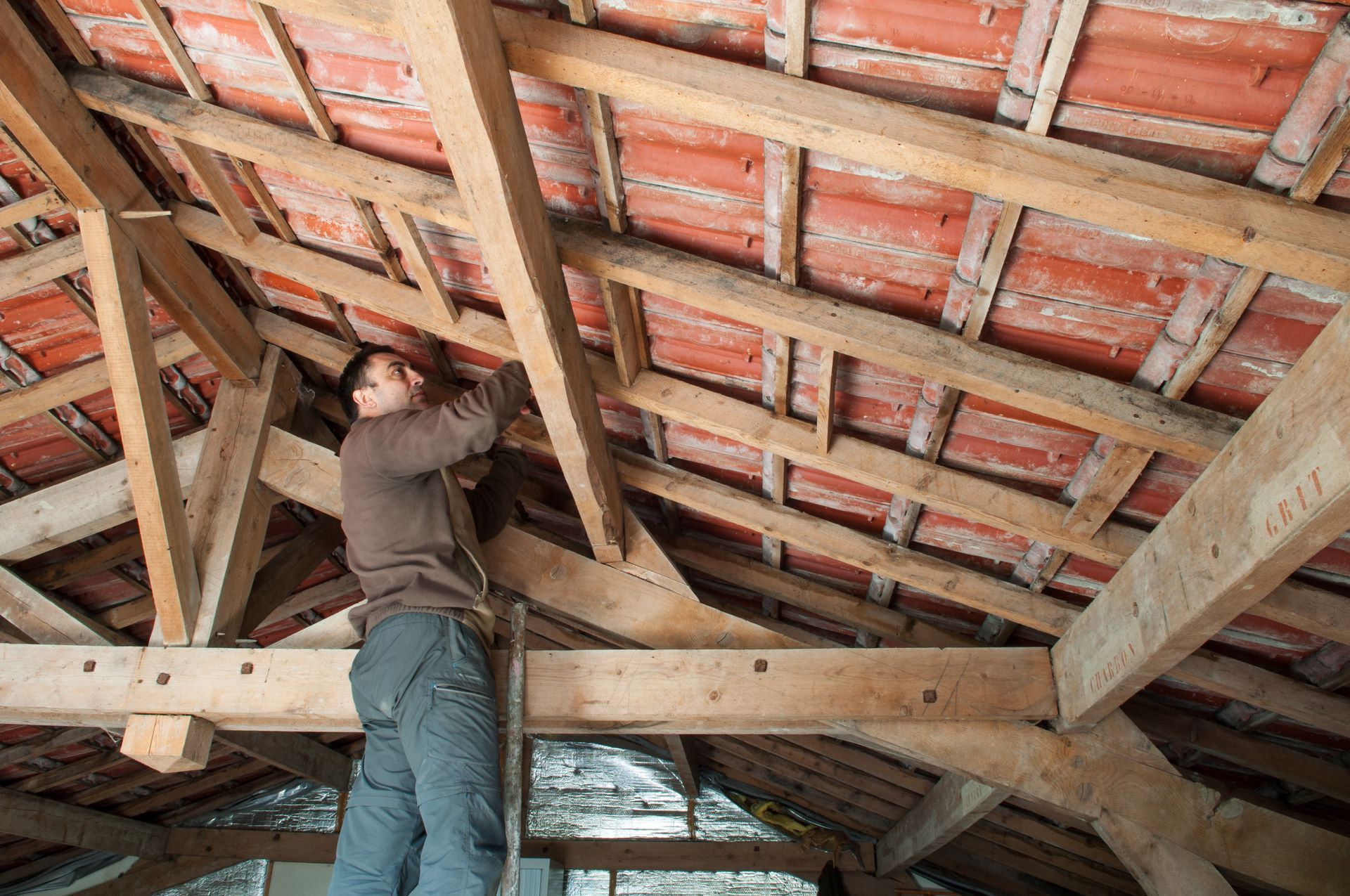 Un homme travaille sur une charpente de toit en bois, on aperçoit des tuiles rouges.