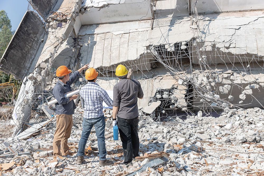Drei Bauarbeiter mit Schutzhelmen untersuchen ein eingestürztes Betongebäude.