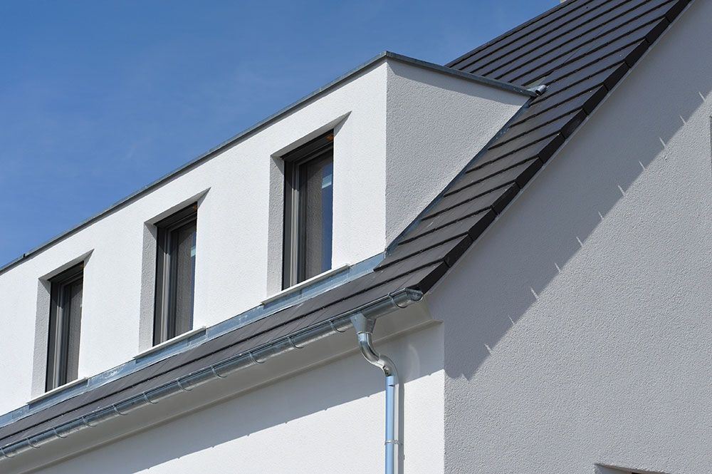 Weißes Haus mit dunklem Dach und drei Fenstern unter blauem Himmel.