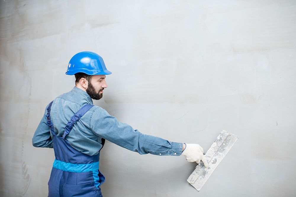 Ein Bauarbeiter in blauem Overall und Schutzhelm glättet mit einer Kelle Putz an einer Wand.