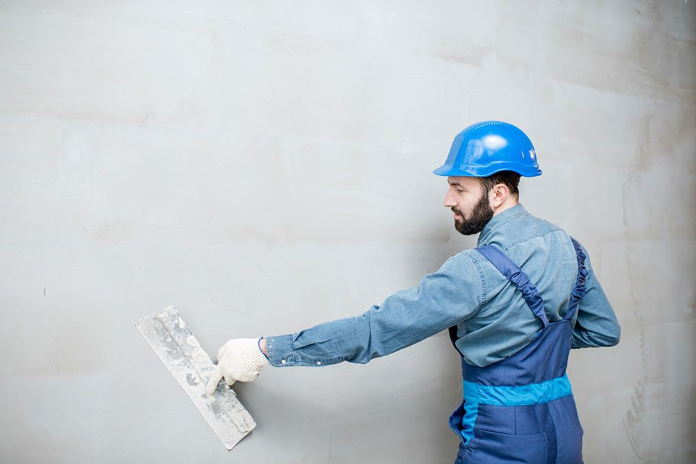 Ein Bauarbeiter in blauem Overall und Schutzhelm glättet mit einer Kelle Putz an einer Wand.