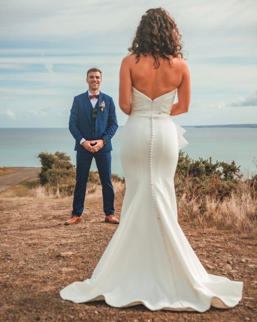 La mariée en robe blanche fait face au marié en costume bleu, l'océan en arrière-plan.