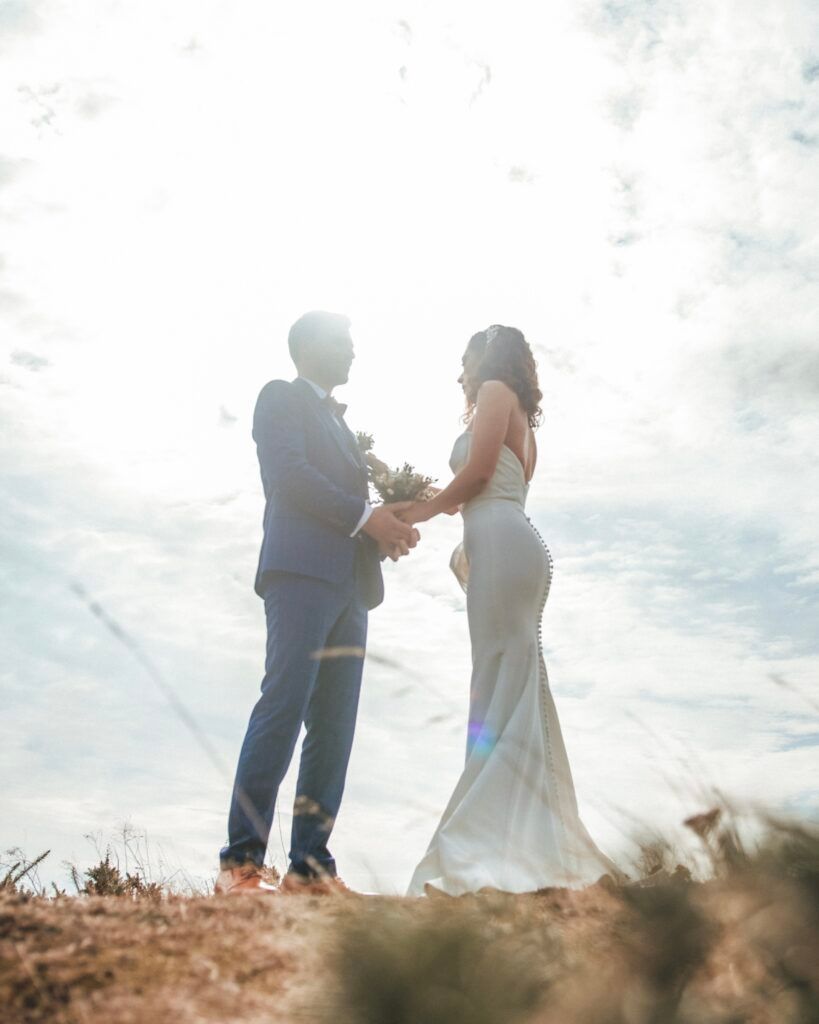 Couple en tenue de mariage se tenant la main, debout sur une colline, se découpant sur fond de soleil éclatant.