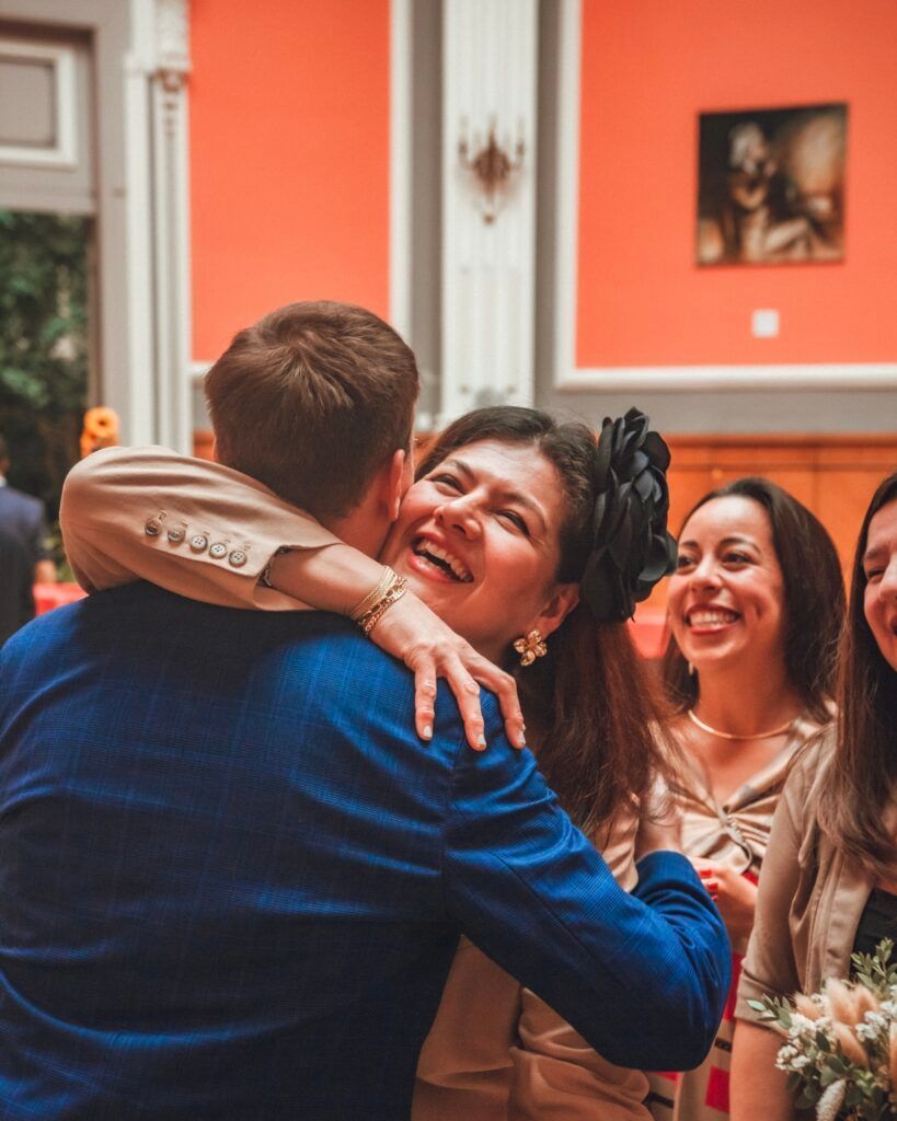 Une femme embrasse un homme, souriant de joie. Deux femmes sourient en arrière-plan. Murs orange vif, intérieur.