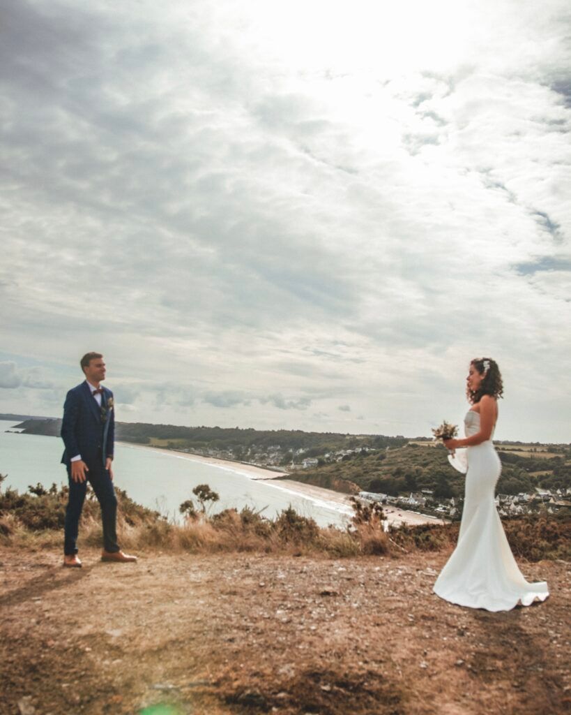 Le marié en costume bleu et la mariée en robe blanche sur une falaise surplombant une plage. Ciel nuageux.