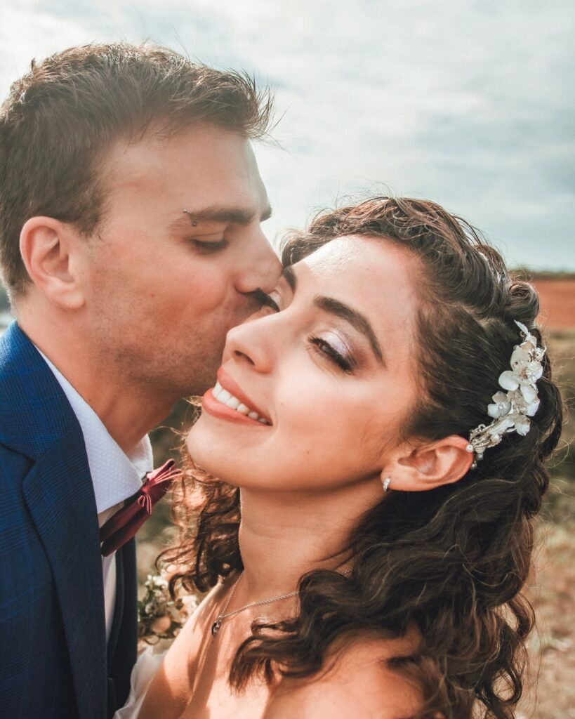 Une mariée souriante avec une coiffe florale est embrassée sur la joue par son marié ; à l'extérieur.