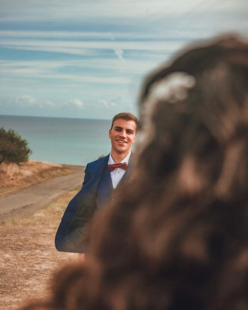 Un homme en costume bleu sourit, face à la caméra sur une route avec la mer en arrière-plan.