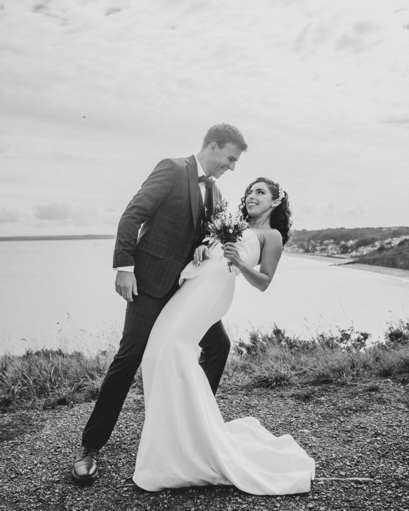 Le marié plonge la mariée sur une falaise surplombant l'océan ; ils rient, vêtus de leur tenue de mariage.