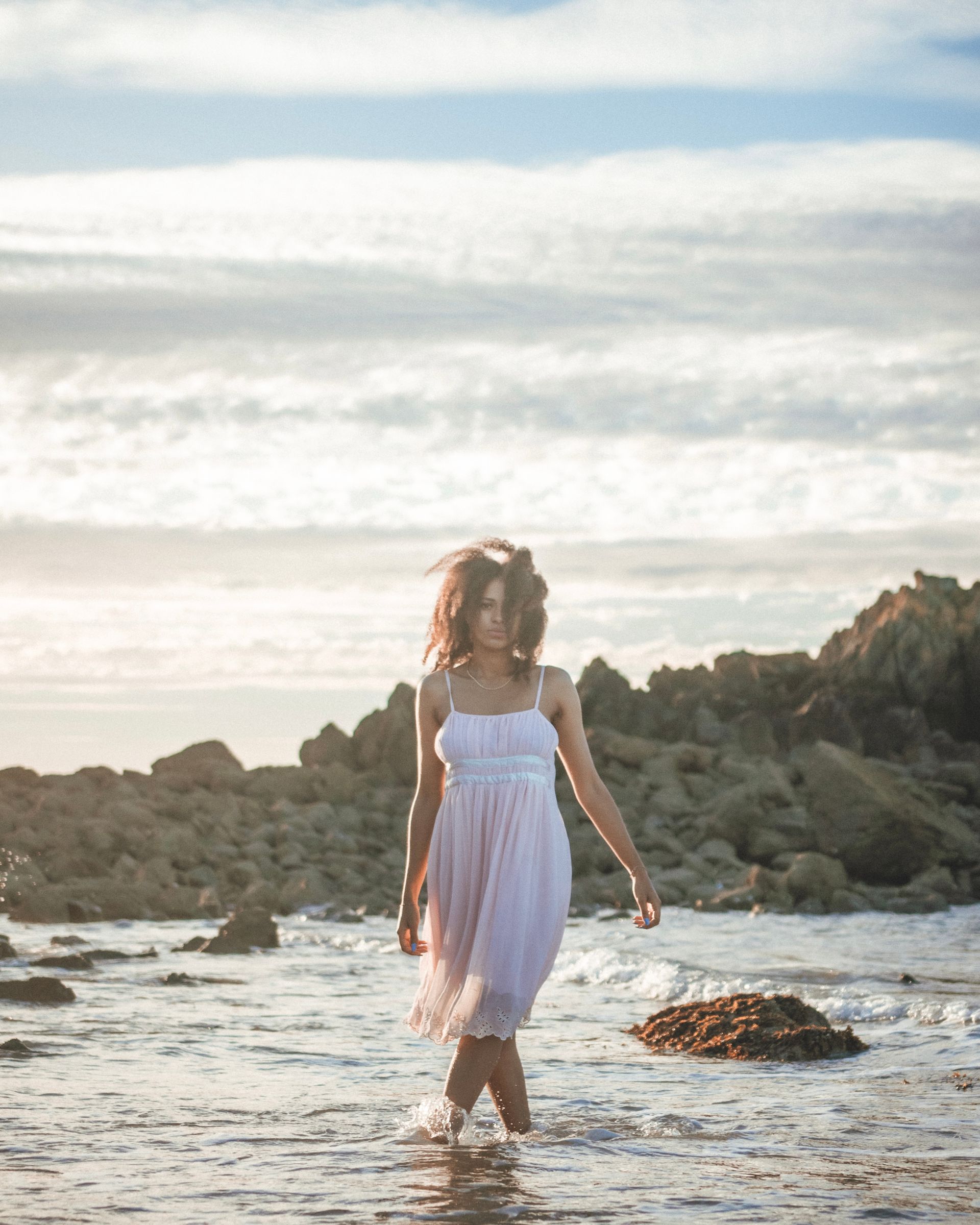Femme en robe blanche pataugeant dans l'océan, éclairé par le soleil, les rochers et le ciel nuageux.